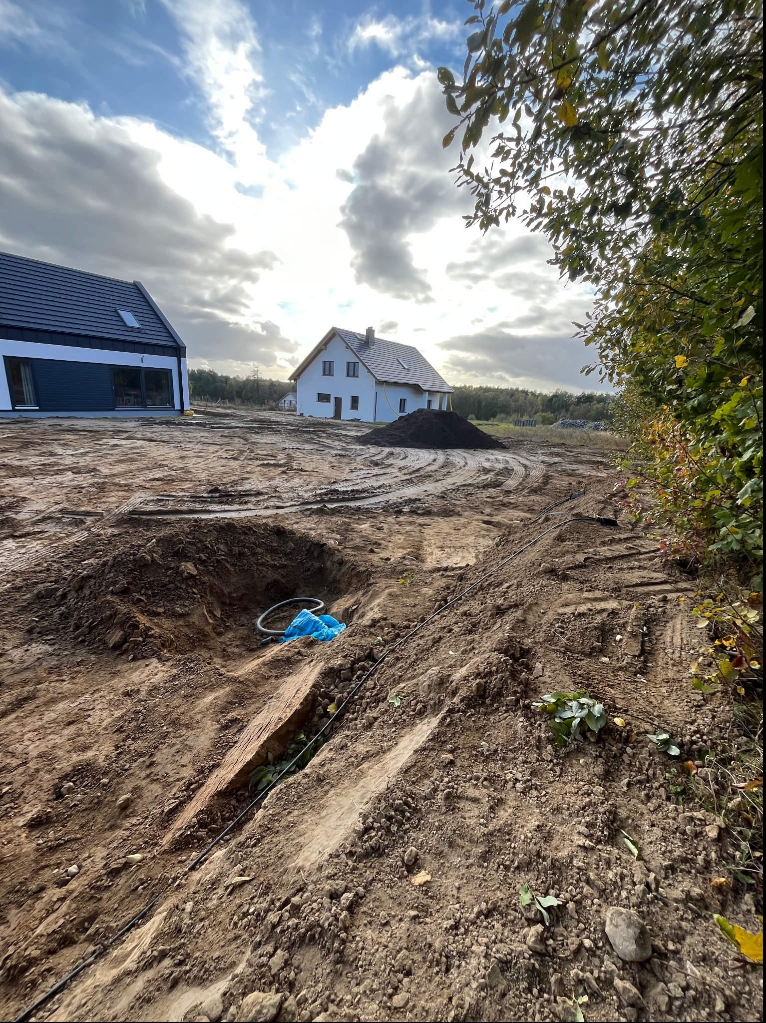 Świeżo wykopany dół w ziemi z niebieską folią i czarnym wężem w środku, w tle dwa domy w stanie surowym otwartym, jeden biały z ciemnym dachem, drugi czarno-biały z ciemnym dachem, ślady opon...