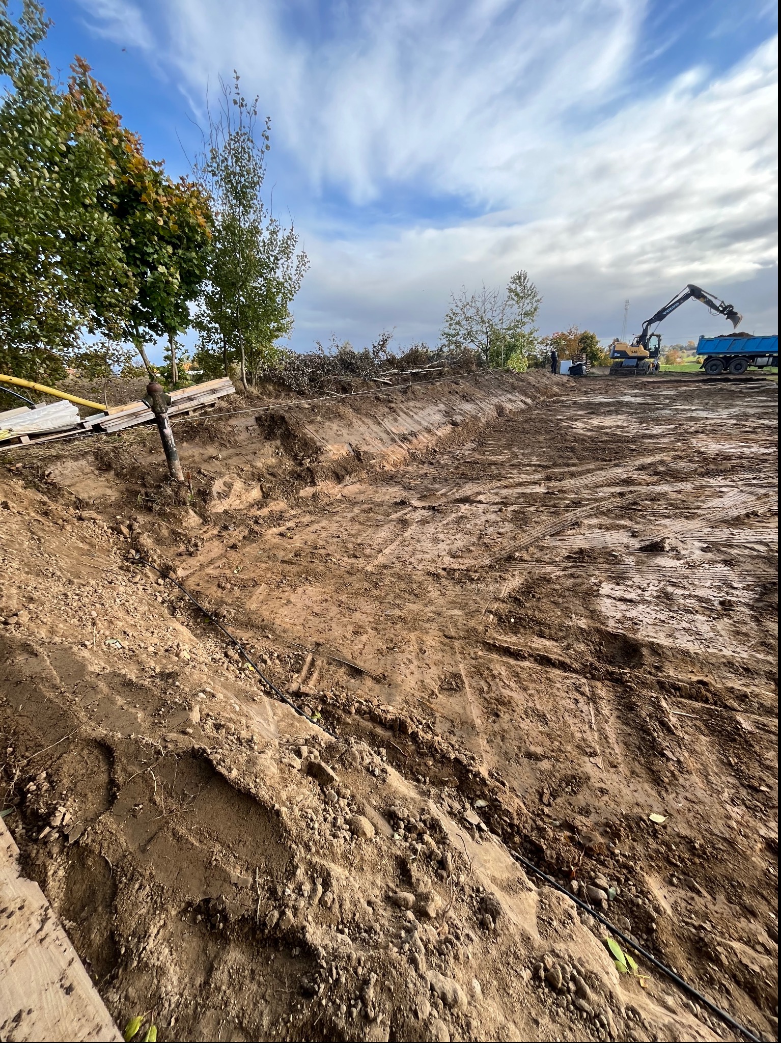 Szerokie ujęcie wykopu w gruncie z widocznymi śladami opon i rurą na dnie, w tle koparka ładująca urobek na ciężarówkę, drzewa i częściowo pochmurne niebo.