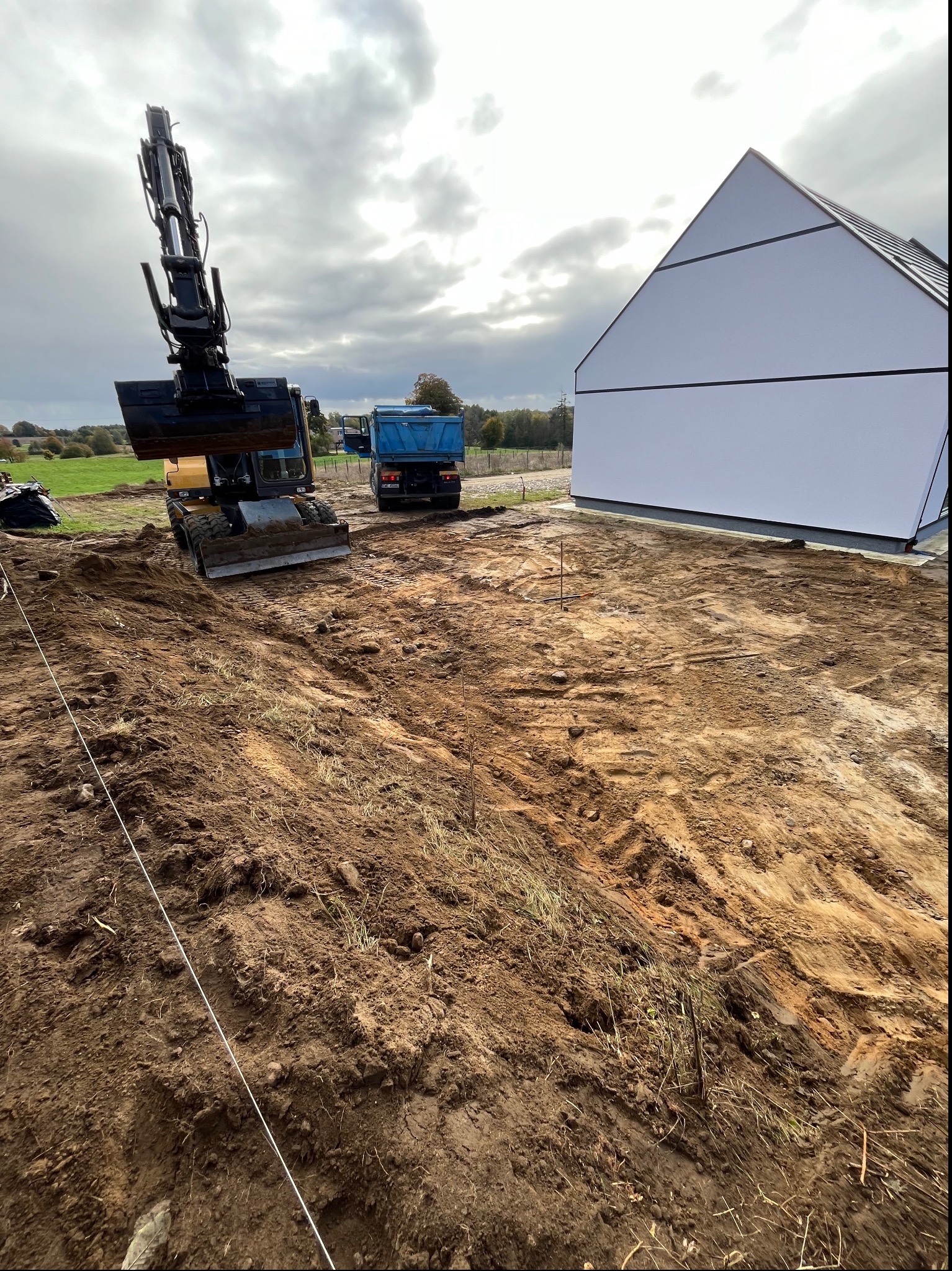 Żółta koparka kołowa i niebieska wywrotka na placu budowy obok białego, nowoczesnego domu z płaskim dachem, teren wytyczony sznurkiem.