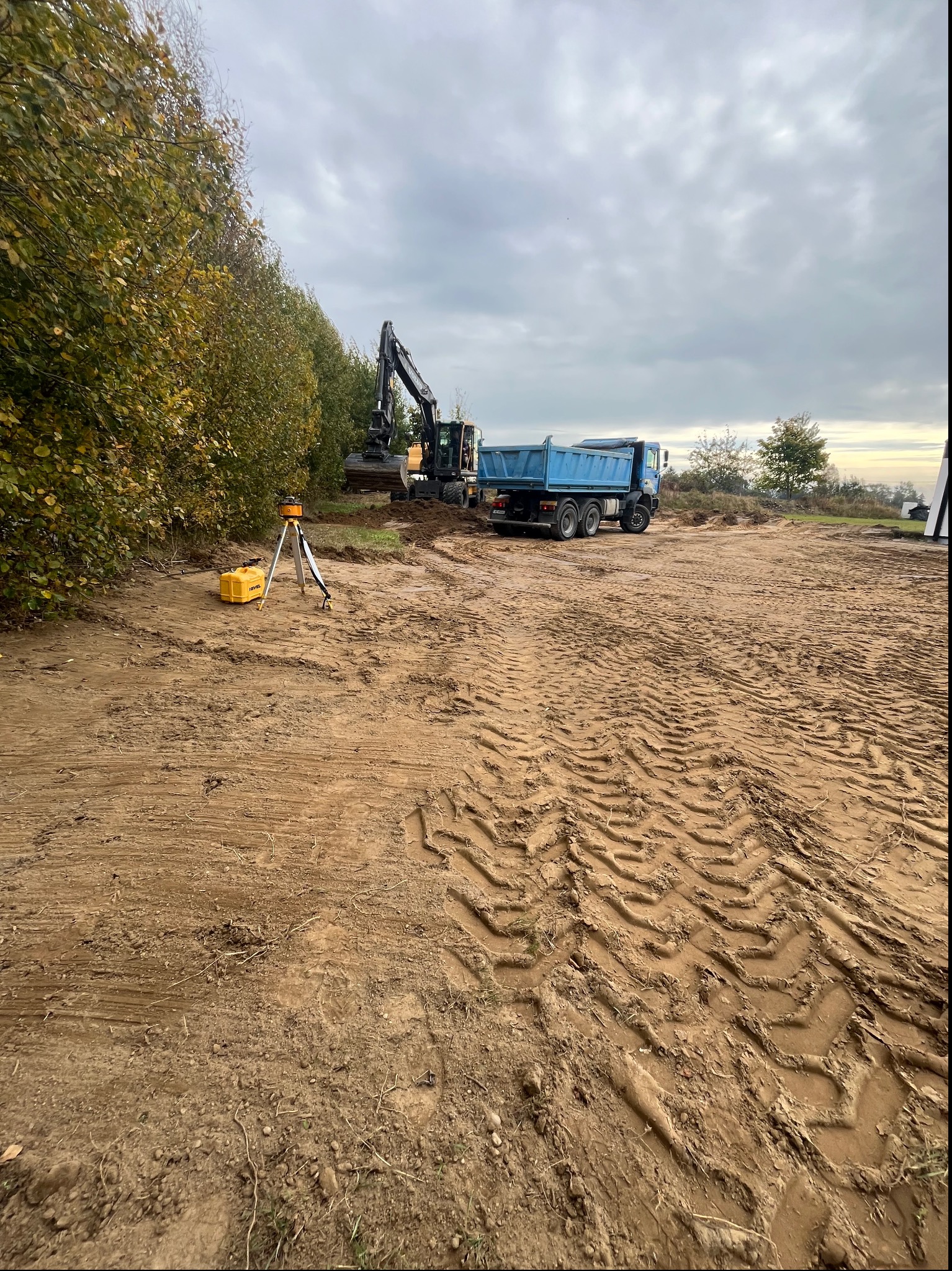 Żółty niwelator laserowy na trójnogu, wykoparka i niebieska wywrotka na placu budowy z błotnistą ziemią i śladami opon, w tle linia drzew i pochmurne niebo.