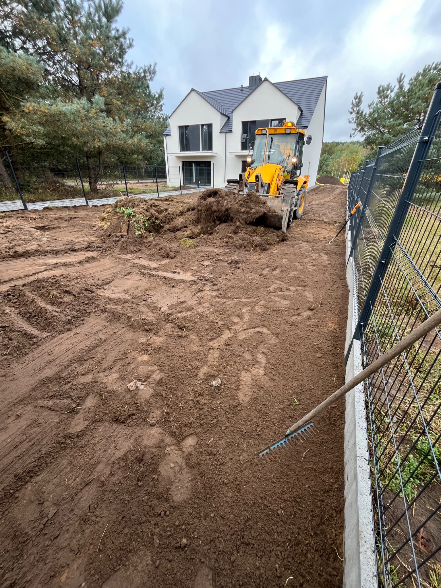 Żółta koparko-ładowarka rozgarnia pryzmę ziemi na działce budowlanej, w tle nowoczesny dom z szarą dachówką i ogrodzenie z siatki.