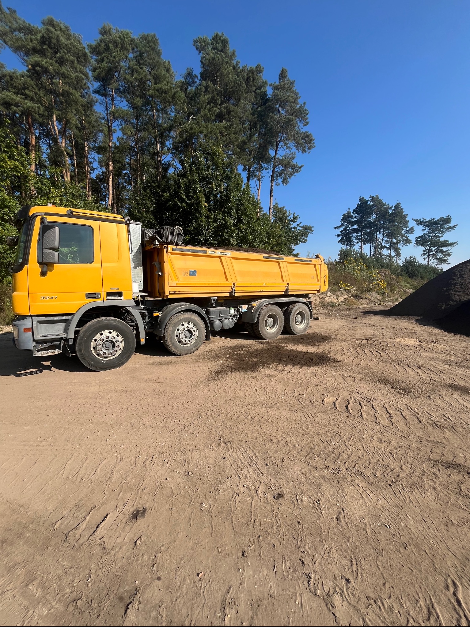 Żółta wywrotka na piaszczystym terenie obok pryzmy ziemi, w tle las iglasty i błękitne niebo.