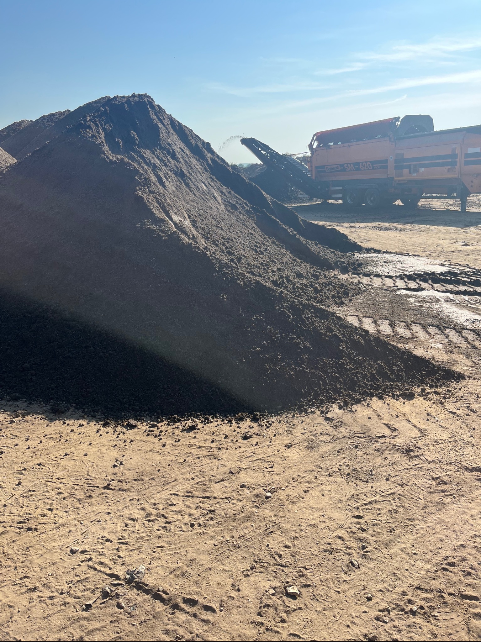 Góra przesiewanej ziemi z pracującym przesiewaczem mobilnym Donestadt SM-620 w tle, widoczne ślady opon na piaszczystym podłożu.