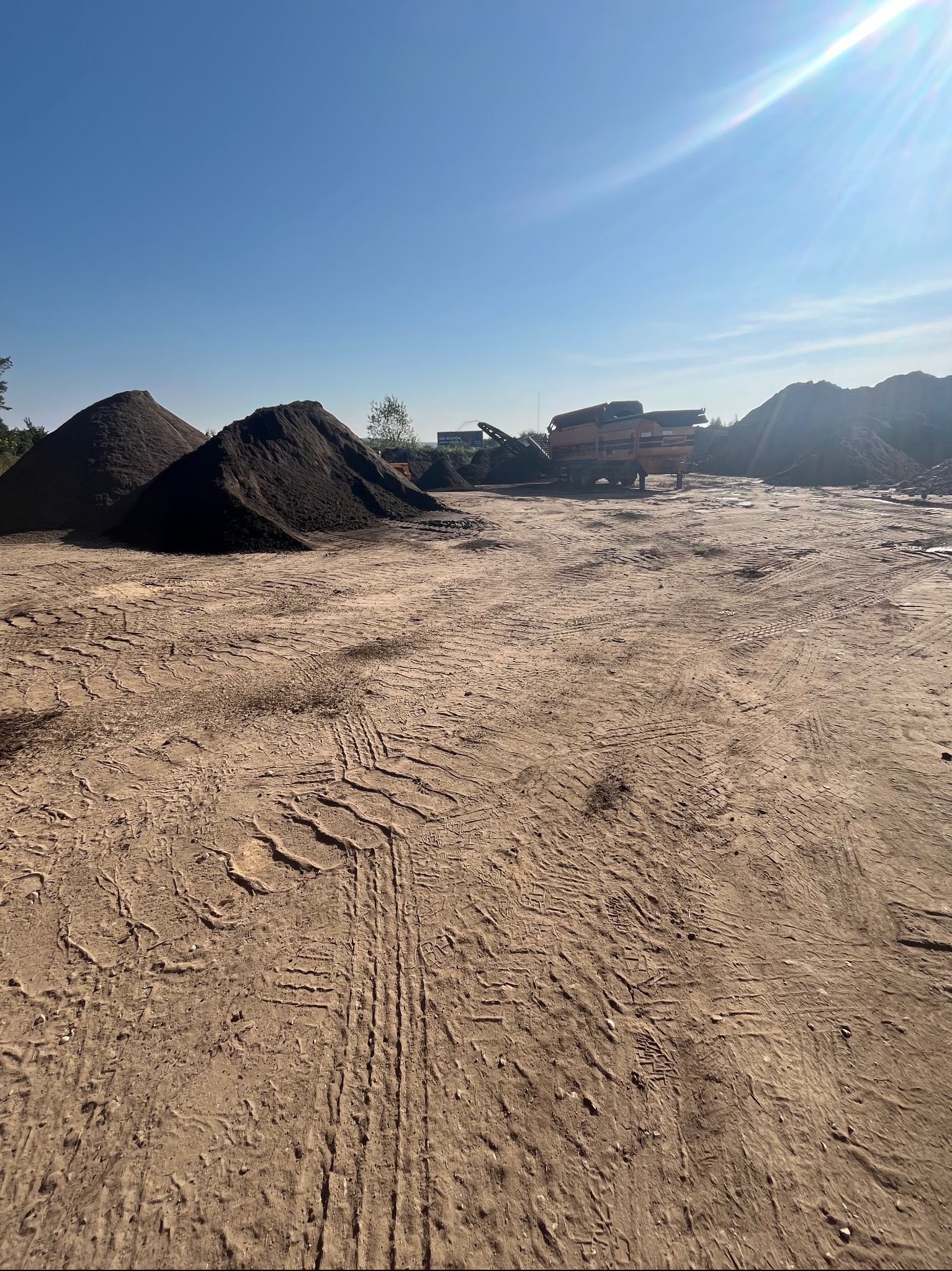 Kielno: Stosy ciemnej ziemi ogrodowej i maszyna do przesiewania na placu, ślady opon na piaszczystym podłożu, błękitne niebo.