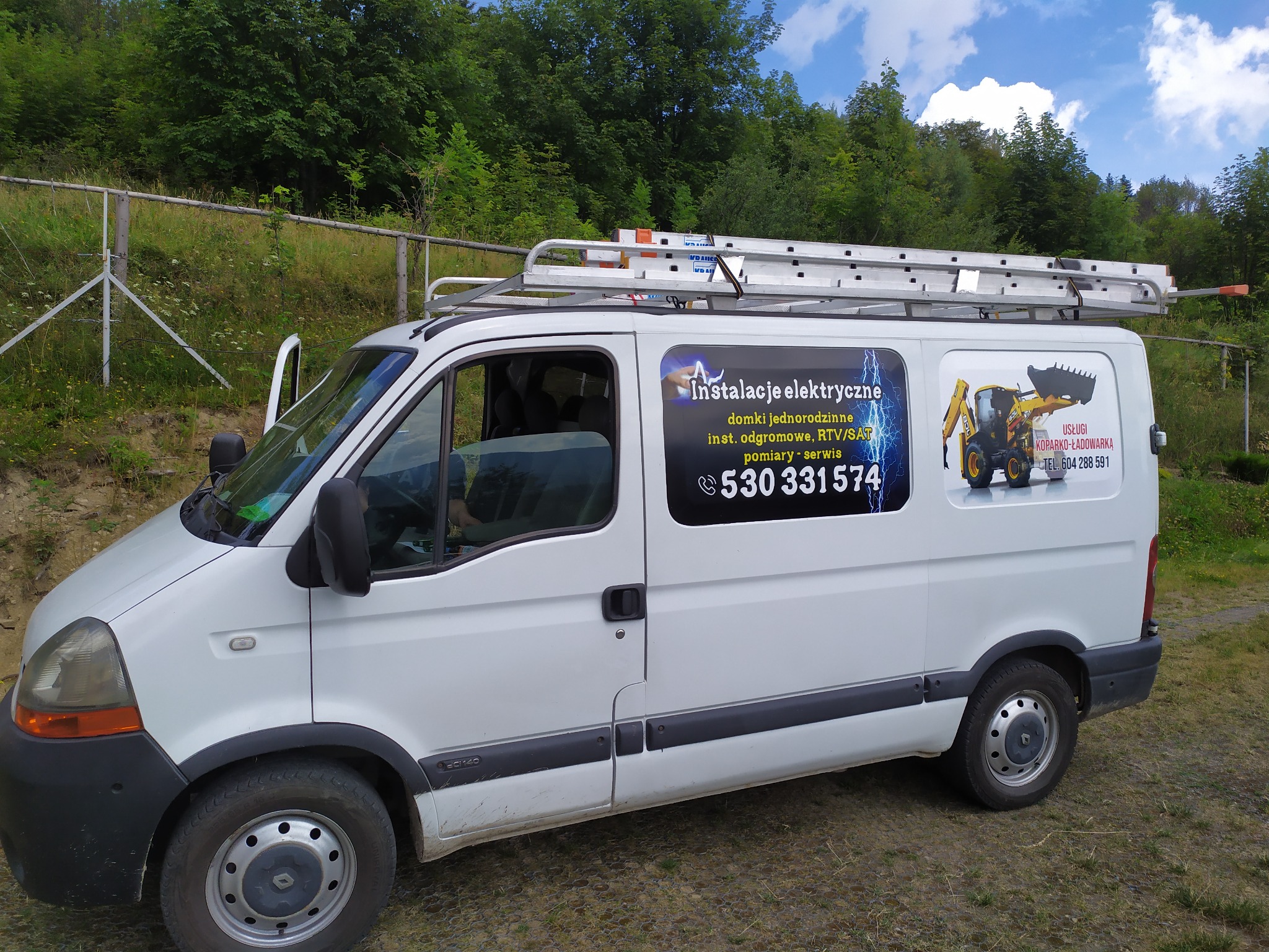 Biały samochód dostawczy z drabiną na dachu, oklejony reklamą usług elektrycznych i koparko-ładowarki, zaparkowany na trawiastym terenie z widokiem na las.