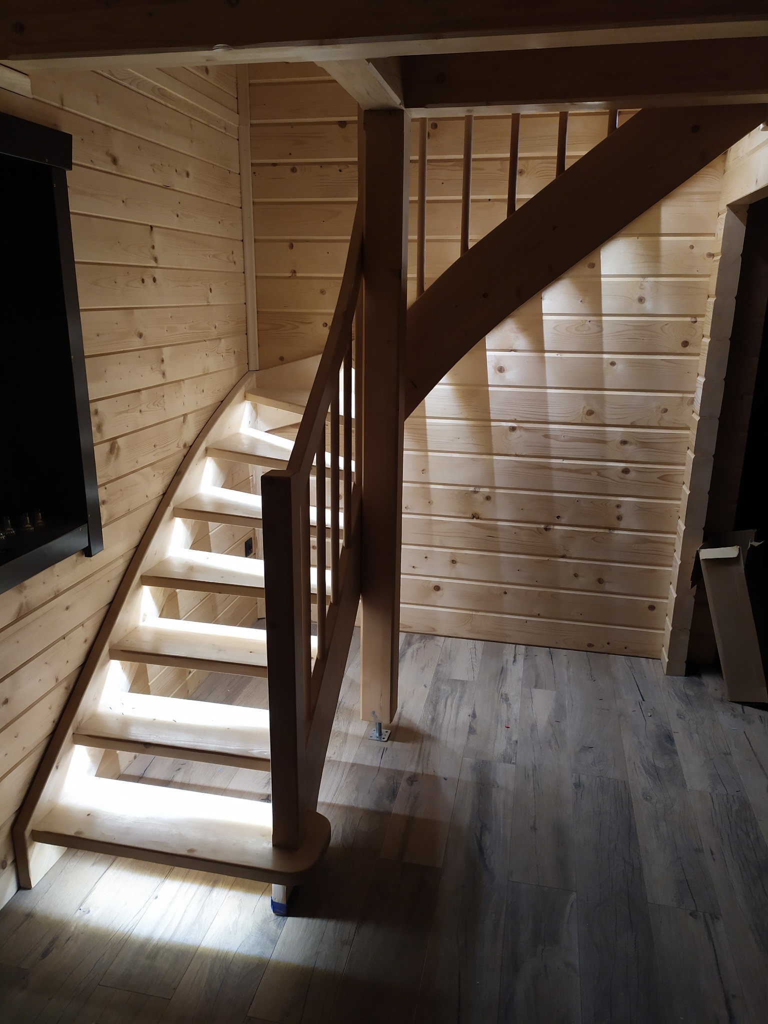 Drewniane schody z podświetleniem LED na każdym stopniu w domu z boazerią, widoczne elementy konstrukcyjne i podłoga imitująca drewno.