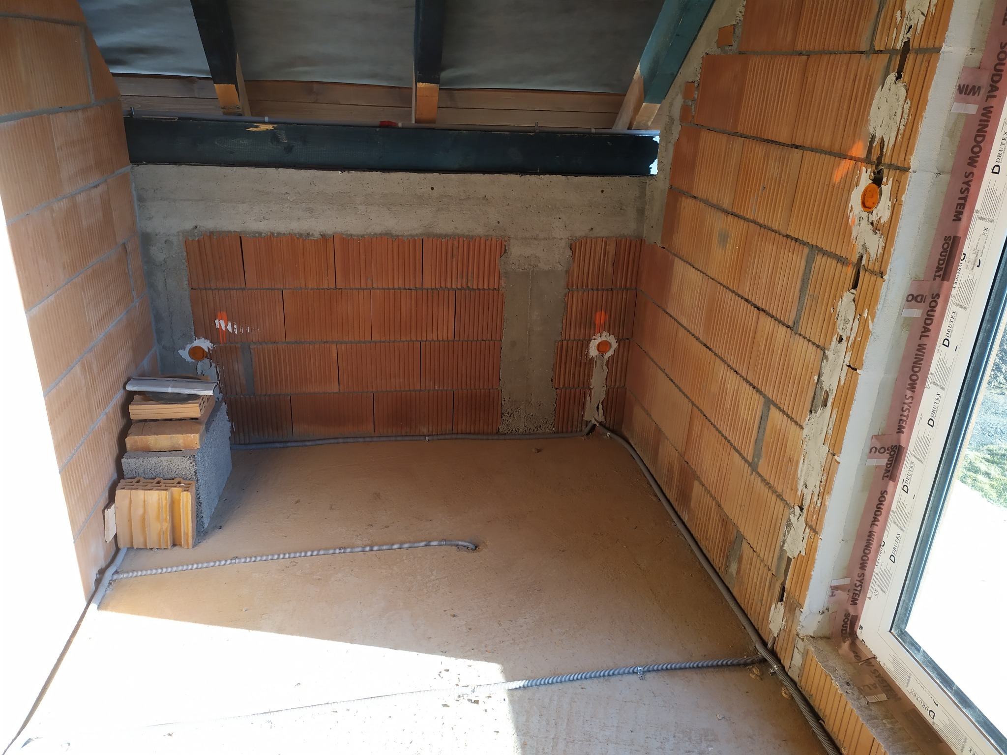 Wnętrze w trakcie budowy z widocznymi przewodami elektrycznymi w peszlach, pomarańczowe puszki instalacyjne w ścianach z cegły ceramicznej, widoczne elementy konstrukcji dachu.