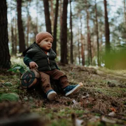 Mały chłopiec w brązowej czapce i zielonej kamizelce siedzi na pniu drzewa w lesie, trzymając dekoracyjny drewniany przedmiot z zielonymi i czerwonymi detalami.