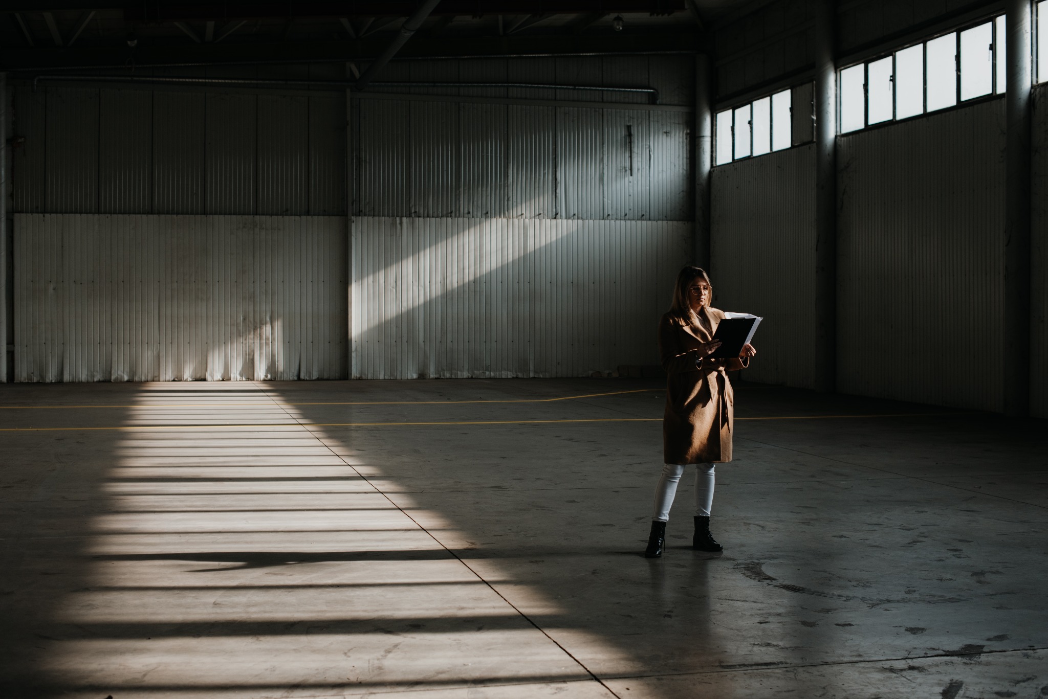Kobieta w płaszczu trzymająca czarny segregator na tle pustej hali z oświetleniem padającym przez okna.