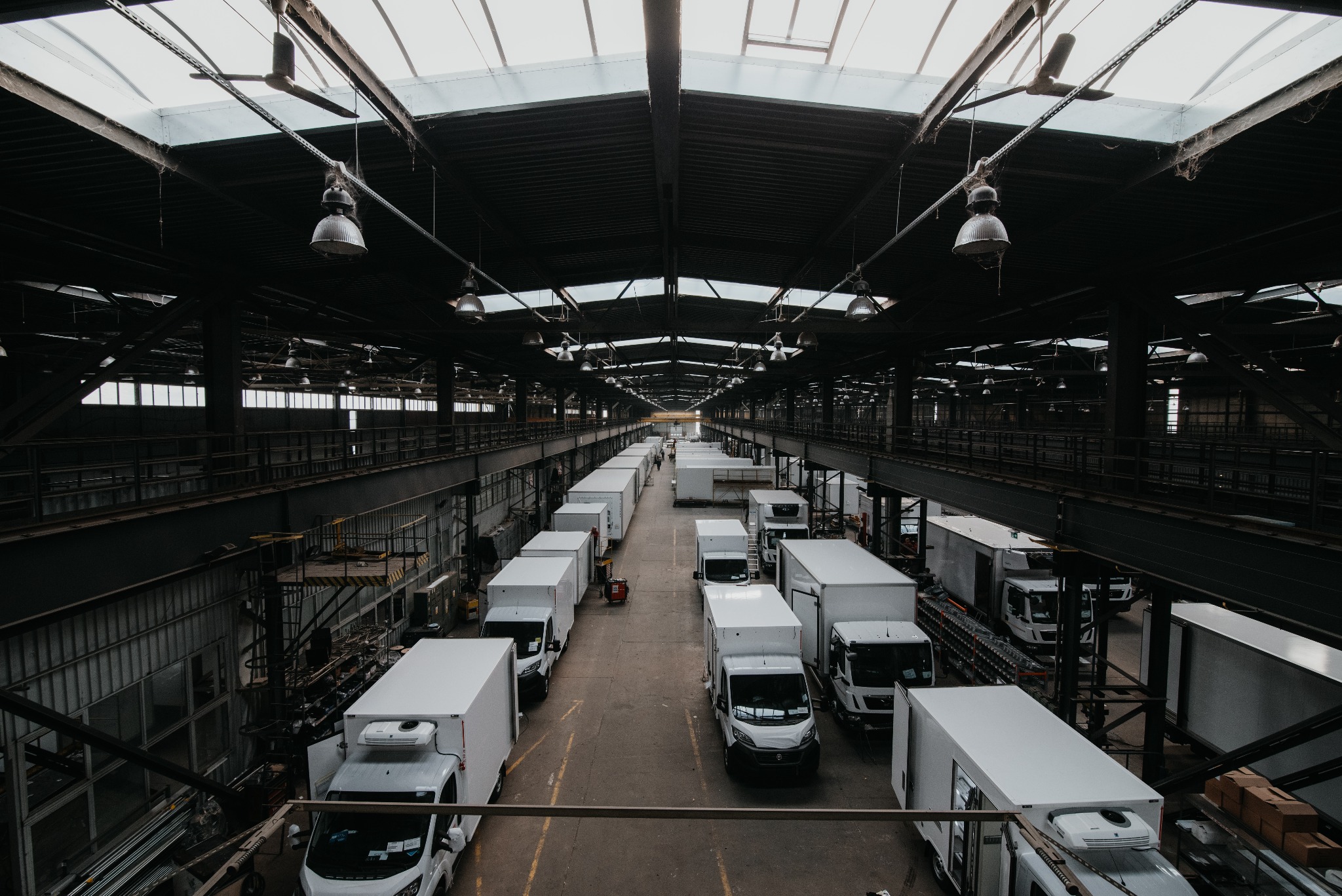 Wysoki kąt widzenia na halę produkcyjną z rzędem białych samochodów dostawczych z chłodniami, oświetlonych przez świetliki w dachu i wiszące lampy.
