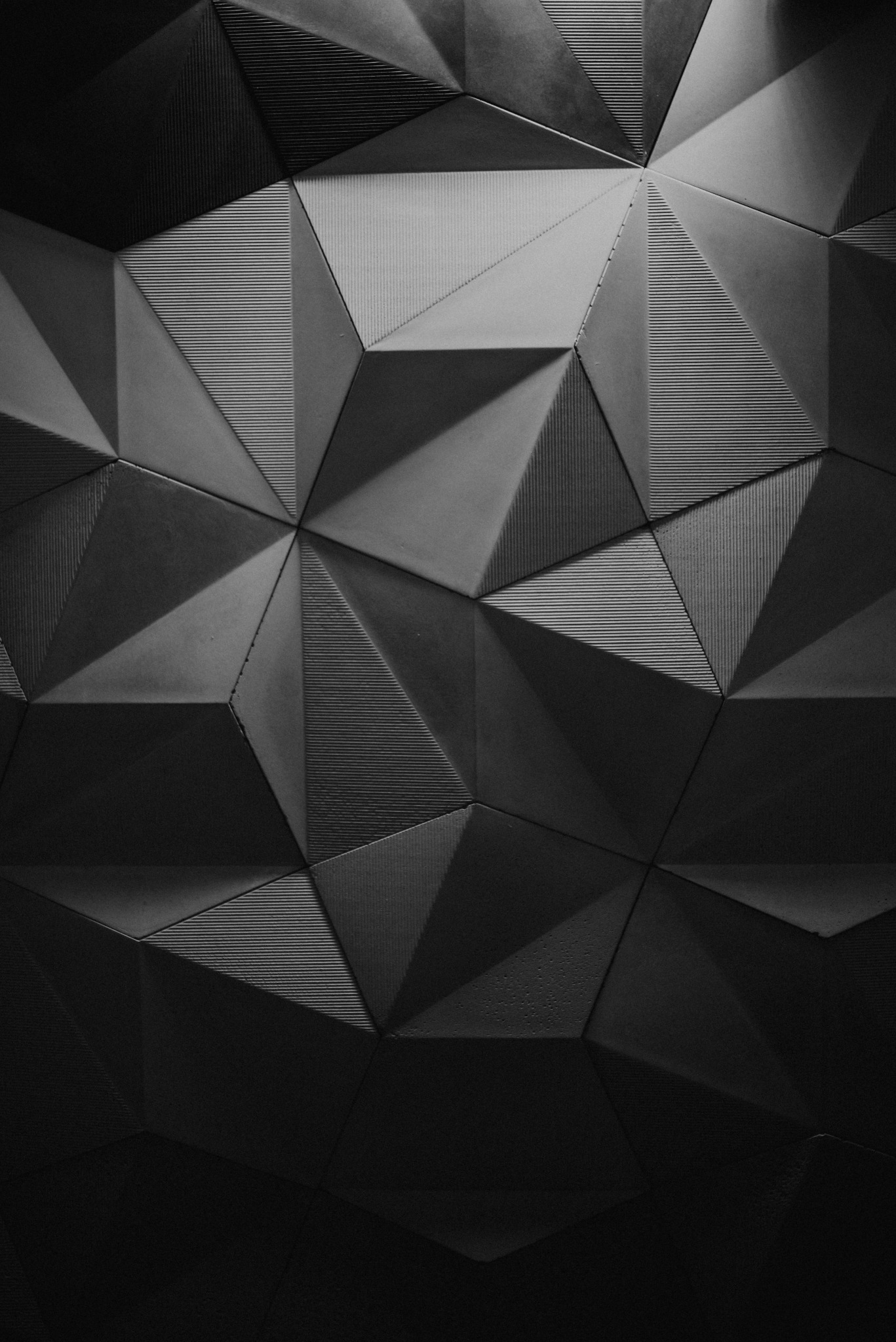 Abstrakcyjna, trójwymiarowa tekstura ściany w odcieniach szarości, złożona z geometrycznych, fasetowanych elementów; niektóre z nich posiadają subtelne, poziome prążkowanie.