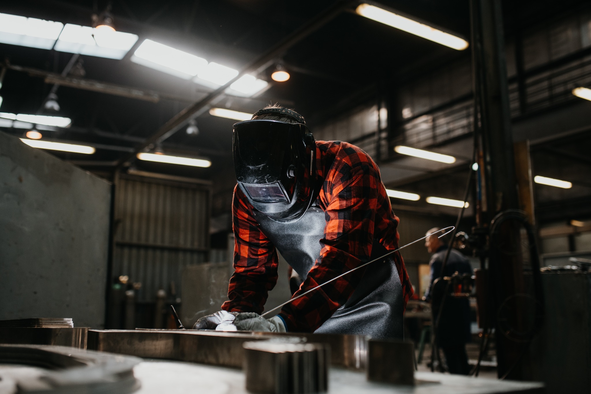 Spawacz w masce ochronnej i czerwonej koszuli w kratę, w trakcie pracy nad metalową konstrukcją w warsztacie, widoczne iskry i elementy konstrukcyjne.