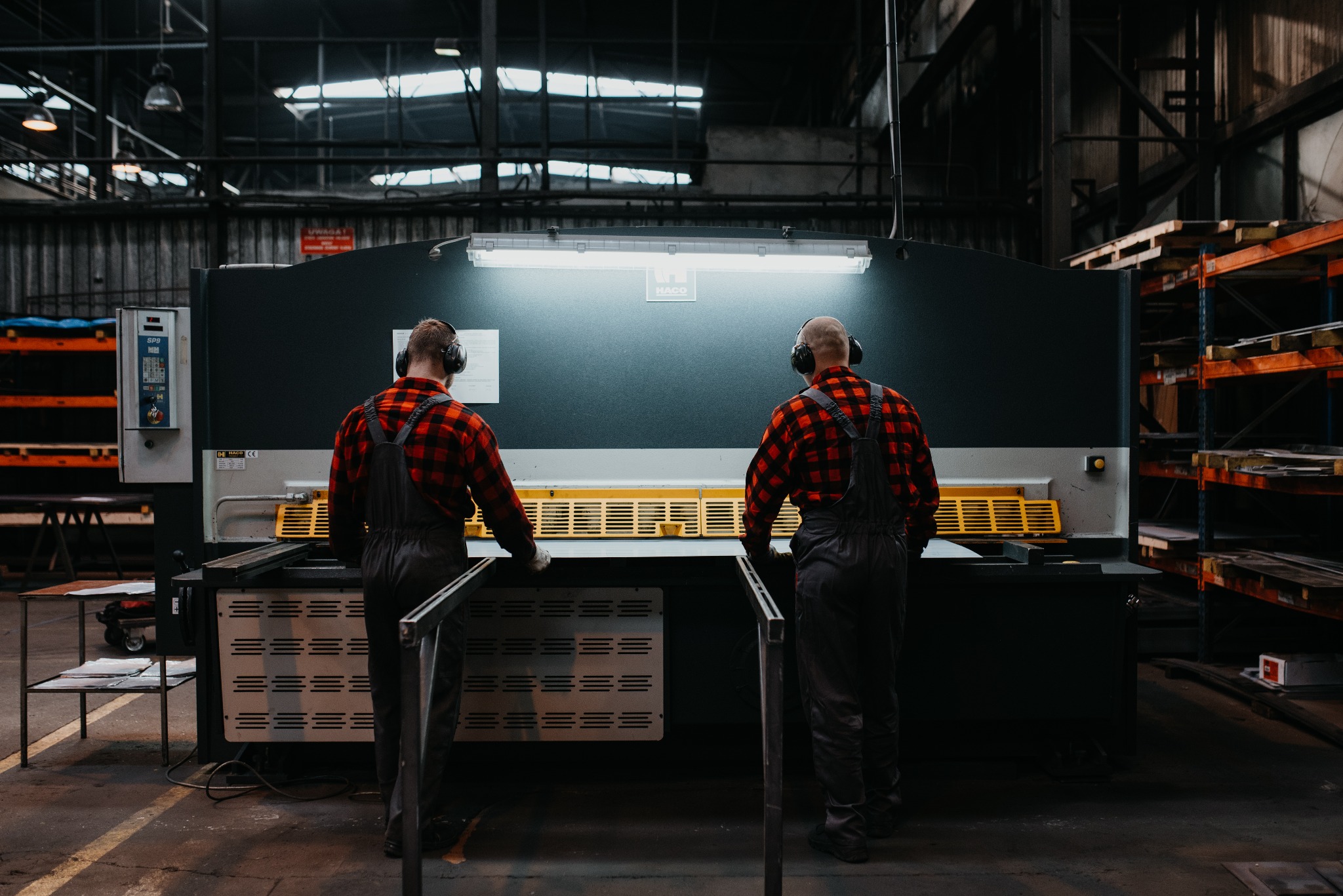 Dwóch pracowników w czerwonych flanelowych koszulach i kombinezonach obsługuje przemysłową gilotynę do cięcia metalu w hali produkcyjnej, obaj mają na uszach słuchawki ochronne.