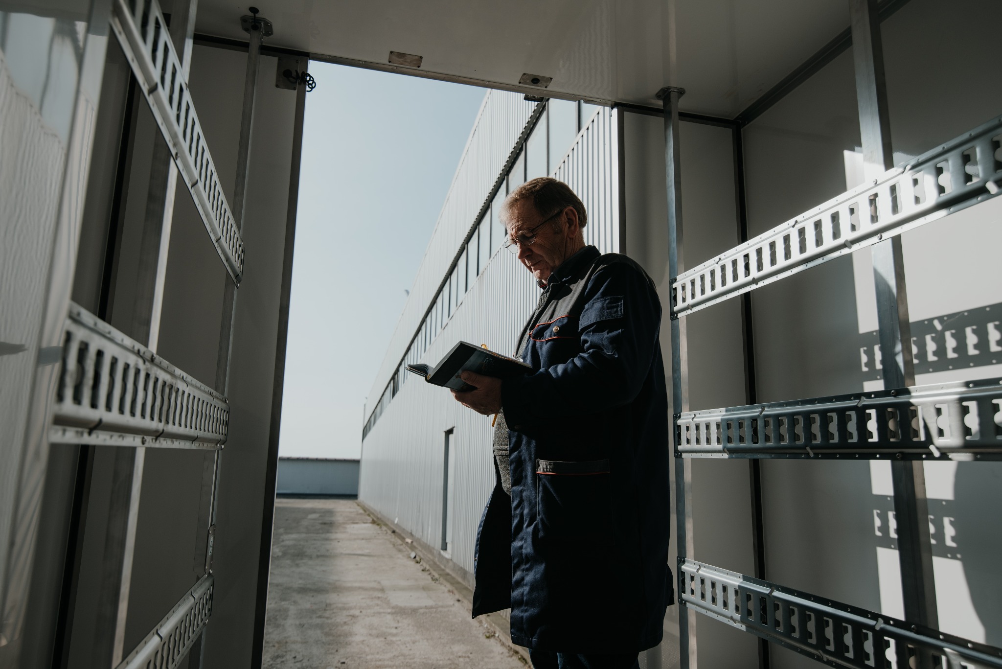 Mężczyzna w okularach i roboczym ubraniu czyta notatnik wewnątrz chłodni z metalowymi szynami do mocowania towarów.