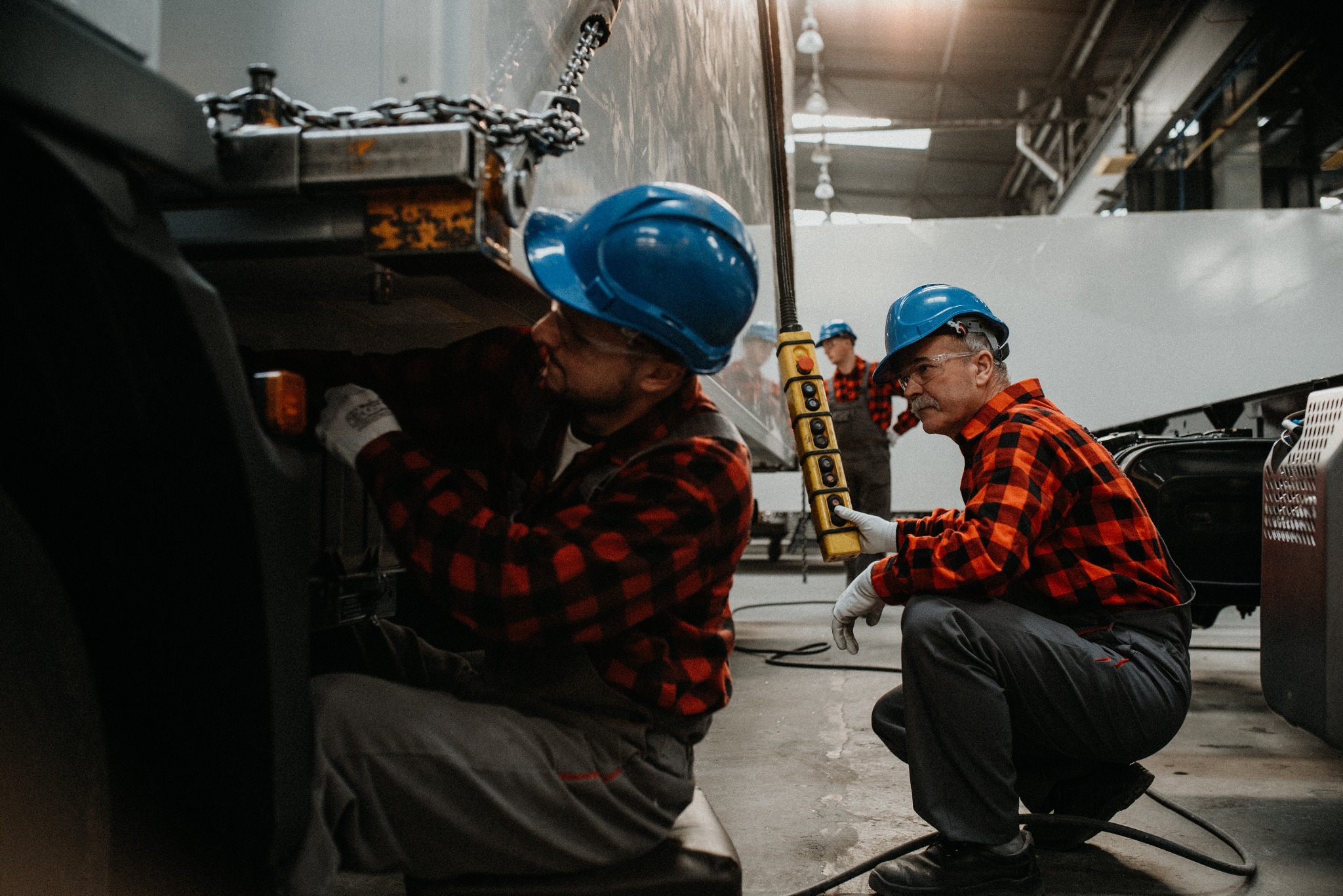 Dwóch mężczyzn w kaskach i flanelowych koszulach pracuje przy montażu ciężarówki w hali, używając dźwigu sterowanego pilotem, z trzecim mężczyzną w tle.
