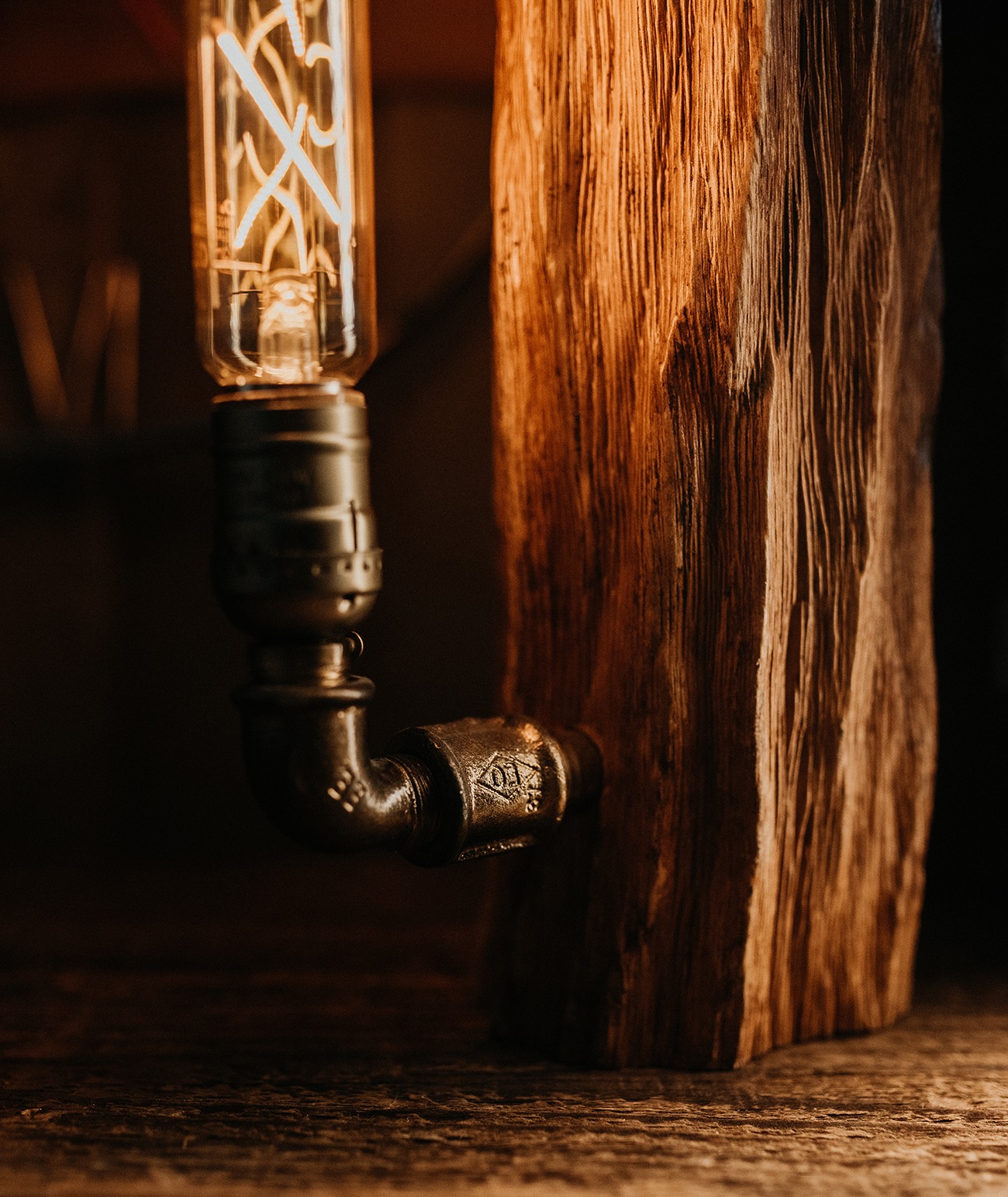 Lampa z żarówką Edisona, zamontowana na drewnianej podstawie z rurami w stylu industrialnym, widoczne detale połączeń rurowych.