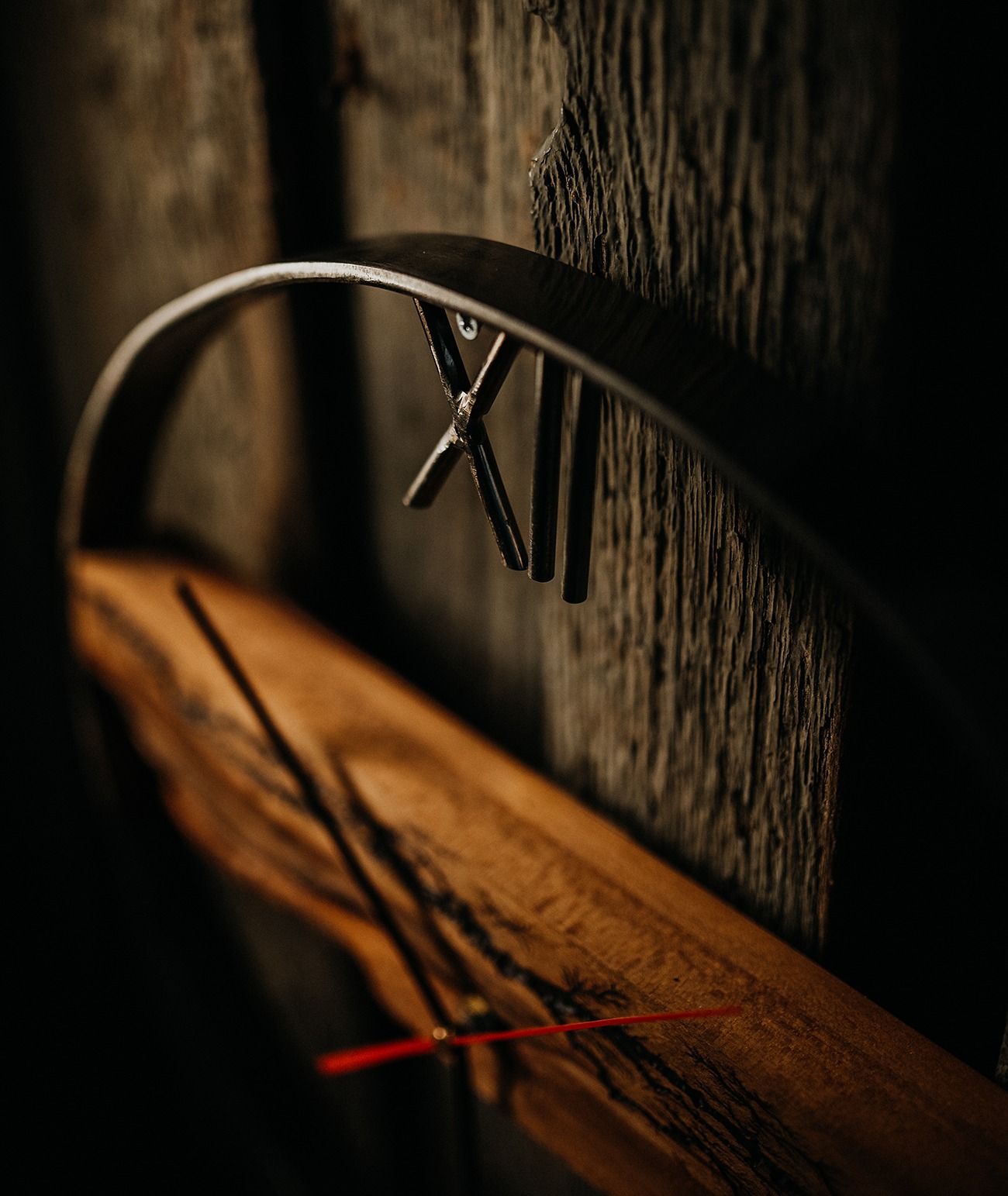 Rustykalny zegar ścienny z drewnianą tarczą i metalową ramą, wskazówki w kolorze czerwonym, zamontowany na ścianie z surowego drewna, ciemne oświetlenie.