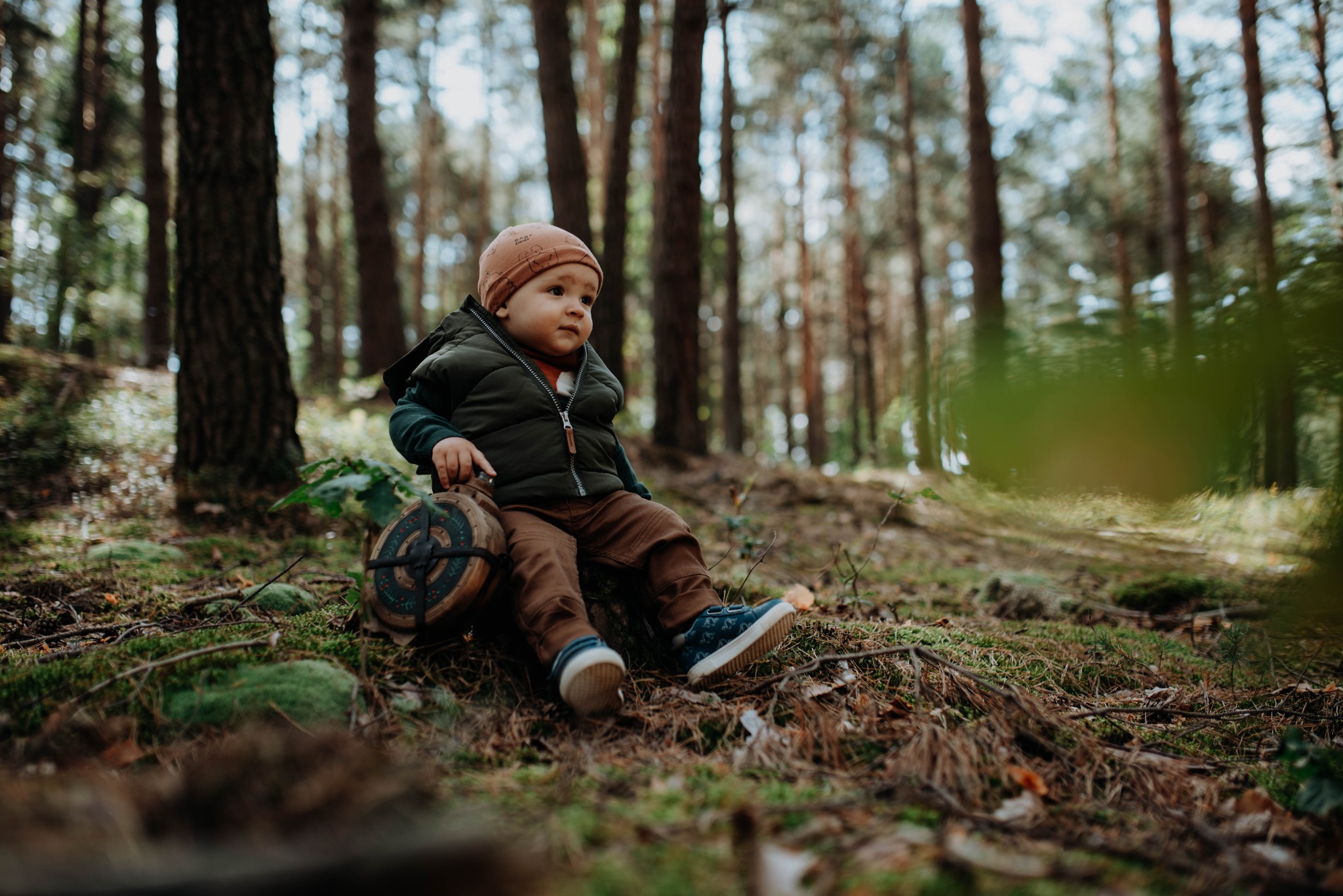 Mały chłopiec w brązowej czapce i zielonej kamizelce siedzi na pniu drzewa w lesie, trzymając dekoracyjny drewniany przedmiot z zielonymi i czerwonymi detalami.
