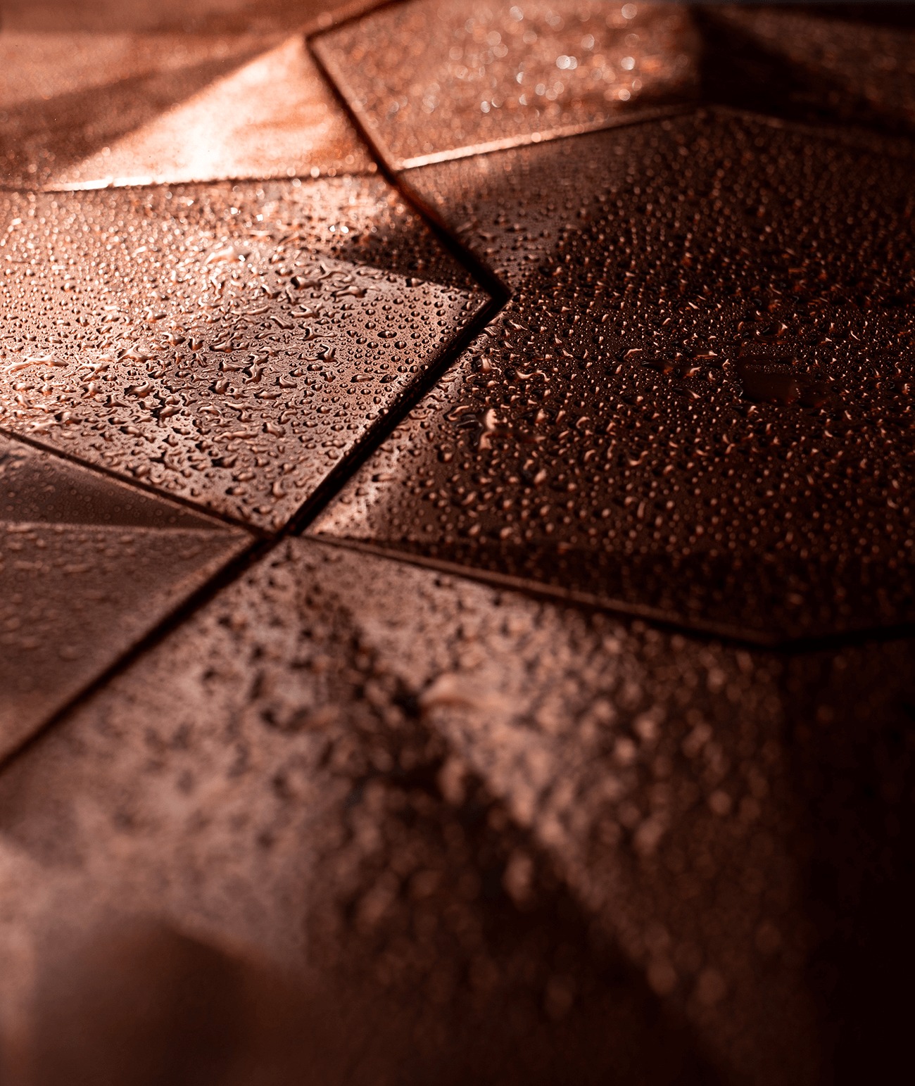 Metalowa powierzchnia pokryta kroplami wody, ułożona z trójkątnych elementów w odcieniach brązu, z bliska i pod kątem.