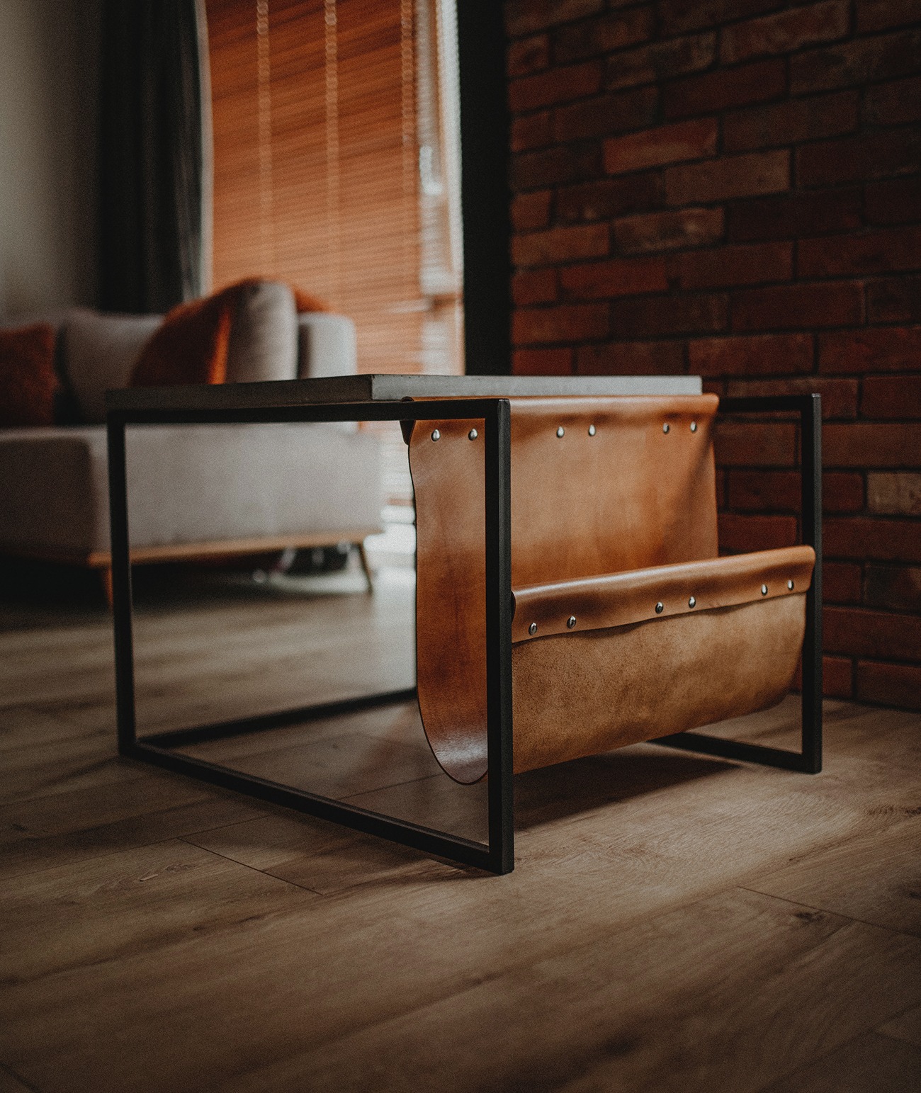 Stolik kawowy z czarną metalową ramą i brązową skórzaną kieszenią na gazety, ustawiony na drewnianej podłodze na tle ceglanej ściany i sofy.