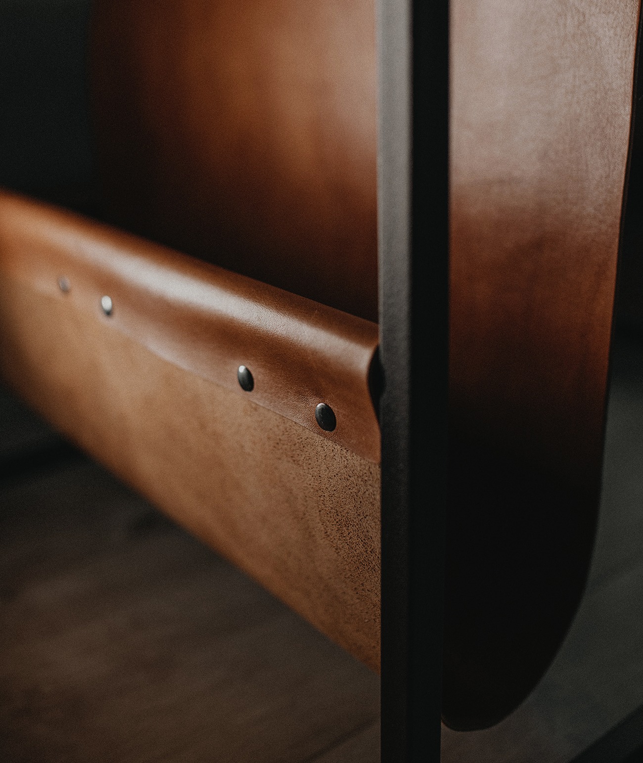 Brązowy skórzany organizer na gazety z nitami, wsparty na czarnej metalowej ramie, z widoczną teksturą skóry i drewnianą podłogą w tle.