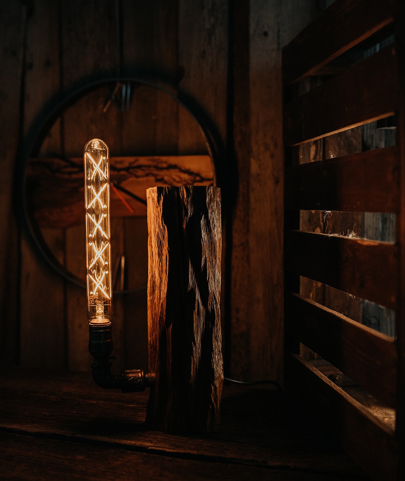 Lampa stojąca z żarówką edisonowską i podstawą z surowego drewna, umieszczona na drewnianym blacie w rustykalnym wnętrzu z okrągłym elementem dekoracyjnym w tle i drewnianą przegrodą z prawej strony.