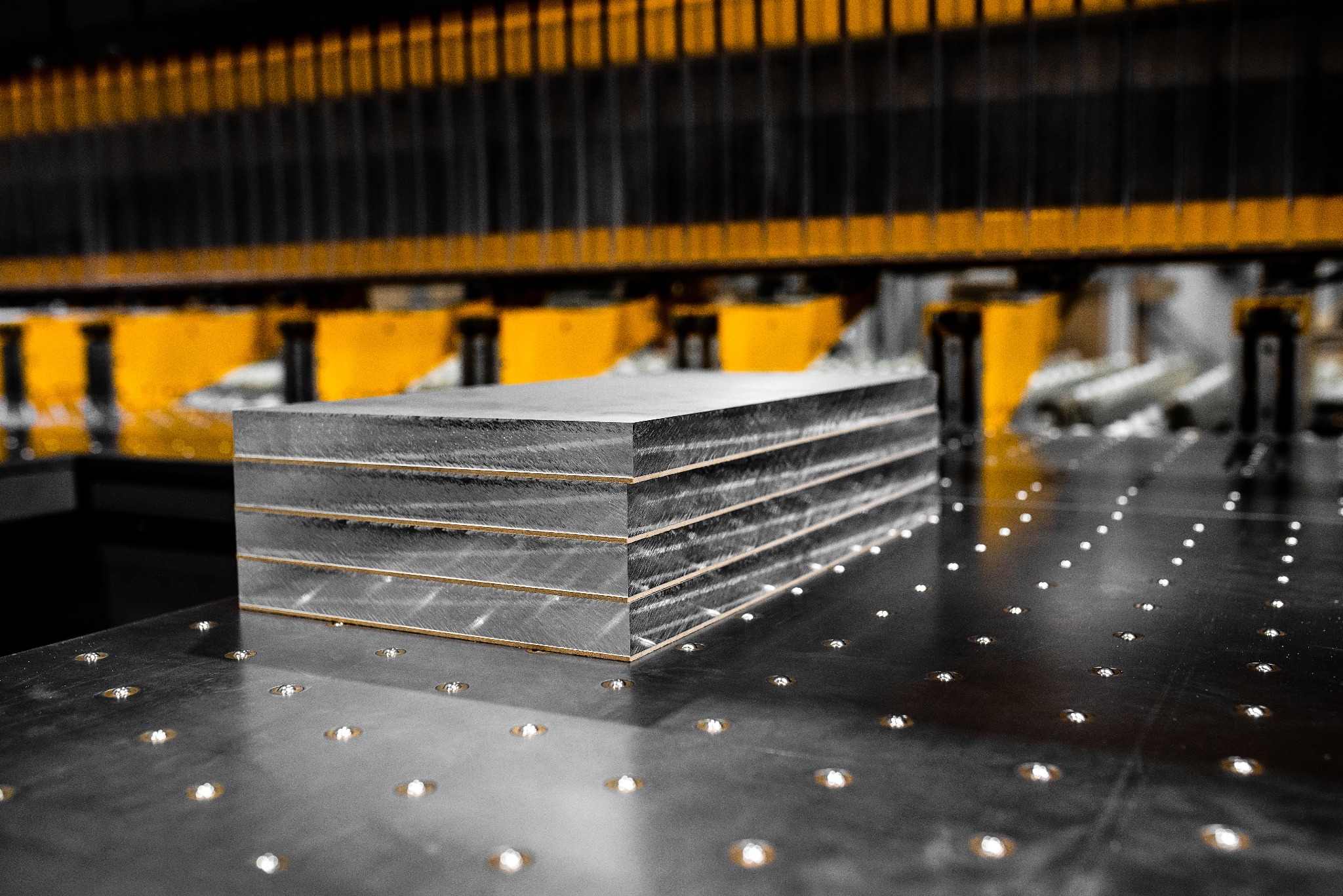 Stos czterech szarych płyt z widocznymi krawędziami na perforowanym stole warsztatowym, w tle żółto-czarna konstrukcja maszyny.