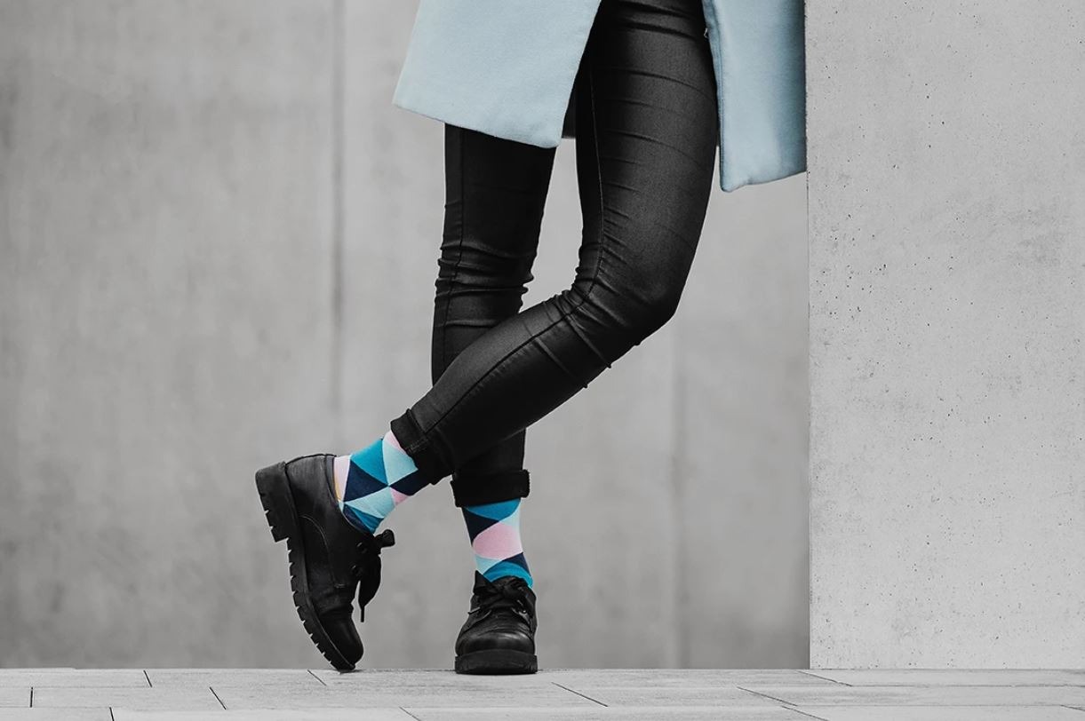 Osoba w czarnych, lakierowanych spodniach i butach, z nogami skrzyżowanymi w kostkach, prezentuje kolorowe skarpetki w geometryczne wzory na tle szarej ściany.