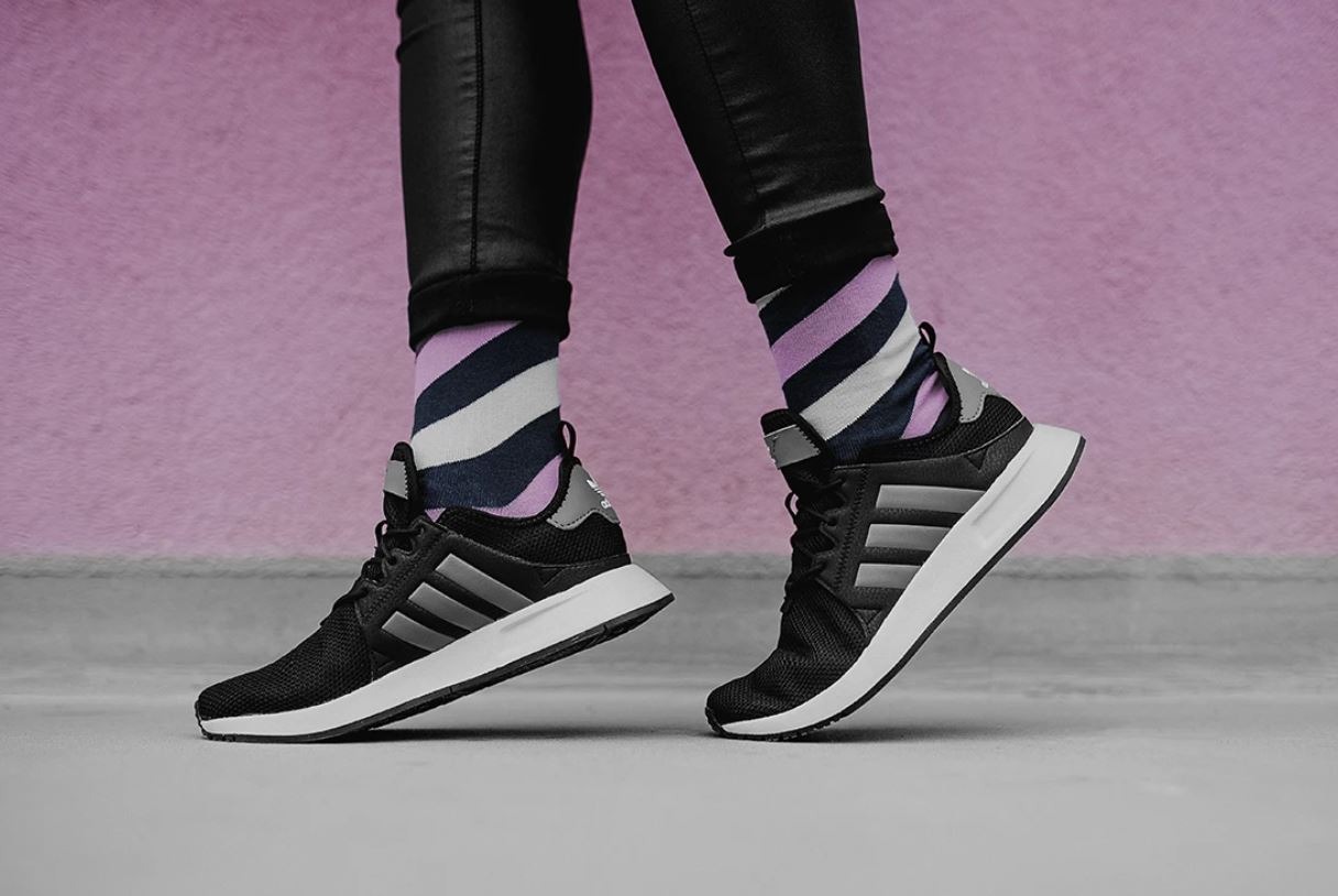 Zbliżenie na stopy w czarnych skórzanych spodniach i czarnych butach sportowych Adidas z szarymi paskami, uzupełnione oryginalnymi skarpetkami w paski fioletu, granatu i bieli na tle fioletowej...