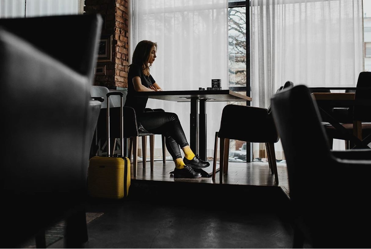 Kobieta siedzi przy stole w kawiarni, obok stoi żółta walizka. Ceglana ściana i okno z zasłonami w tle.