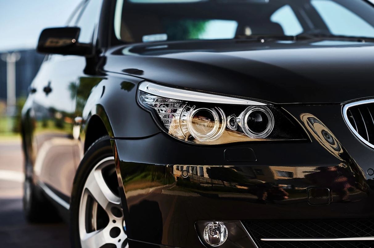 Błyszczący, czarny samochód osobowy, zbliżenie na przedni reflektor i felgę, z widocznymi detalami chromowanymi i odbiciami otoczenia w lakierze.