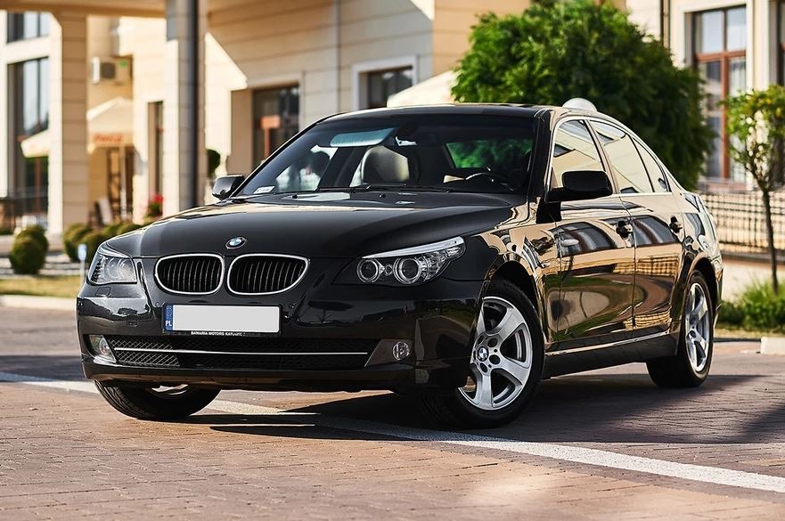 Czarny samochód BMW serii 5, model E60, zaparkowany na brukowanej powierzchni przed budynkiem z kolumnami, w słoneczny dzień.