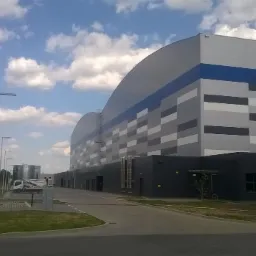 Nowoczesna hala przemysłowa z elewacją w odcieniach szarości i błękitu, widoczna z perspektywy ulicy z elementami infrastruktury miejskiej.