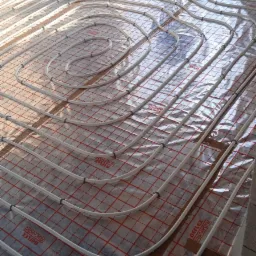 Instalacja ogrzewania podłogowego w surowym stanie, rury grzewcze ułożone na folii z siatką, widoczne okno i ściany z bloczków.