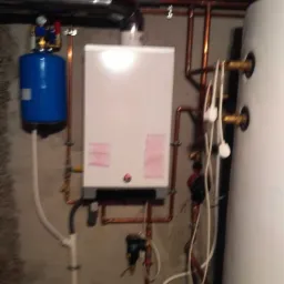 Biały piec z podłączonymi miedzianymi rurami, niebieskim zbiornikiem wyrównawczym i białym zasobnikiem ciepłej wody, zamontowany na szarej, betonowej ścianie w piwnicy.