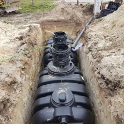 Czarny, zbiornik z tworzywa sztucznego, częściowo zakopany w wykopie ziemnym, widoczne rury i przewody, w tle koparka i budynek mieszkalny.