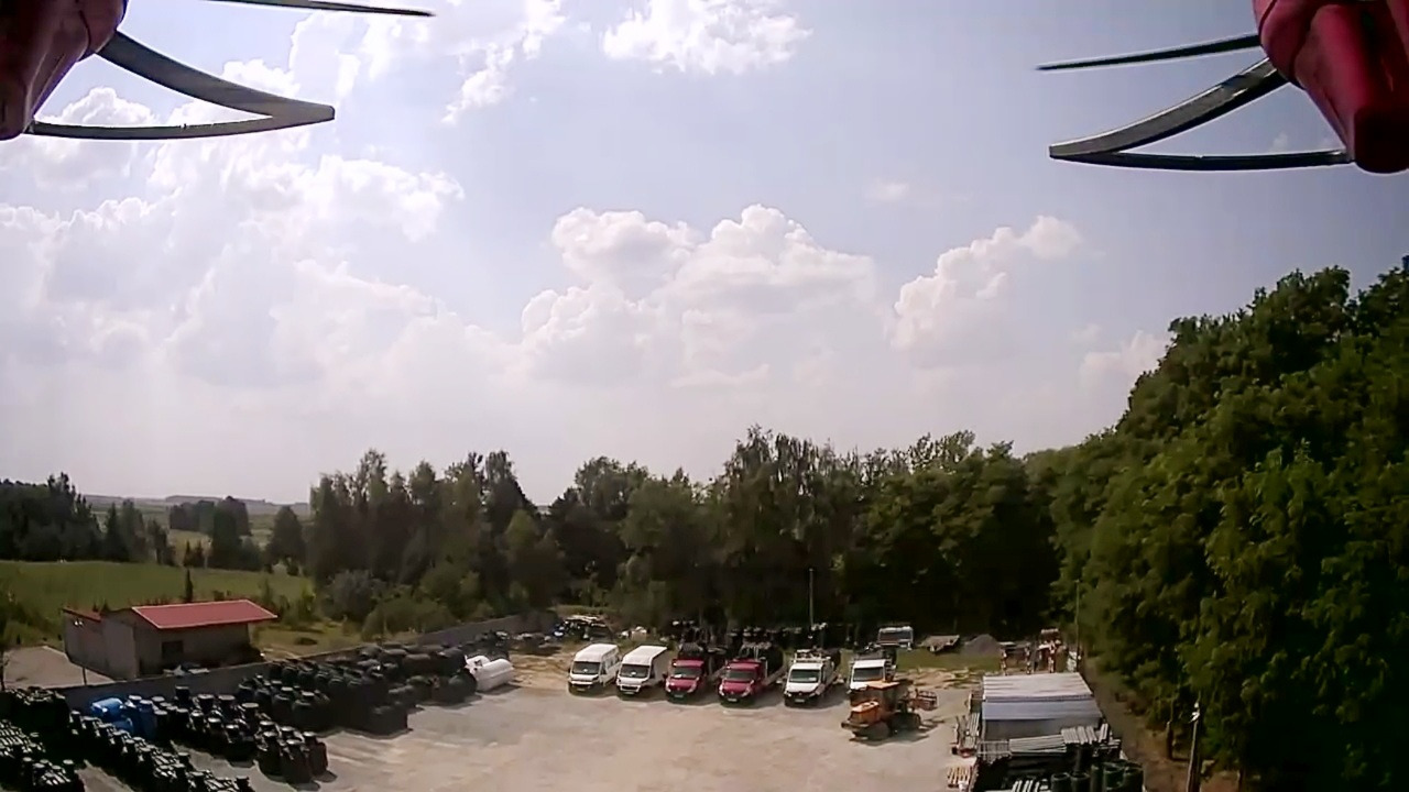 Widok z drona na plac z zapleczem firmy: ułożone rzędy czarnych zbiorników, kilka samochodów dostawczych i ciężarówek, budynek z czerwonym dachem, w tle zieleń drzew i pola uprawne pod błękitnym...