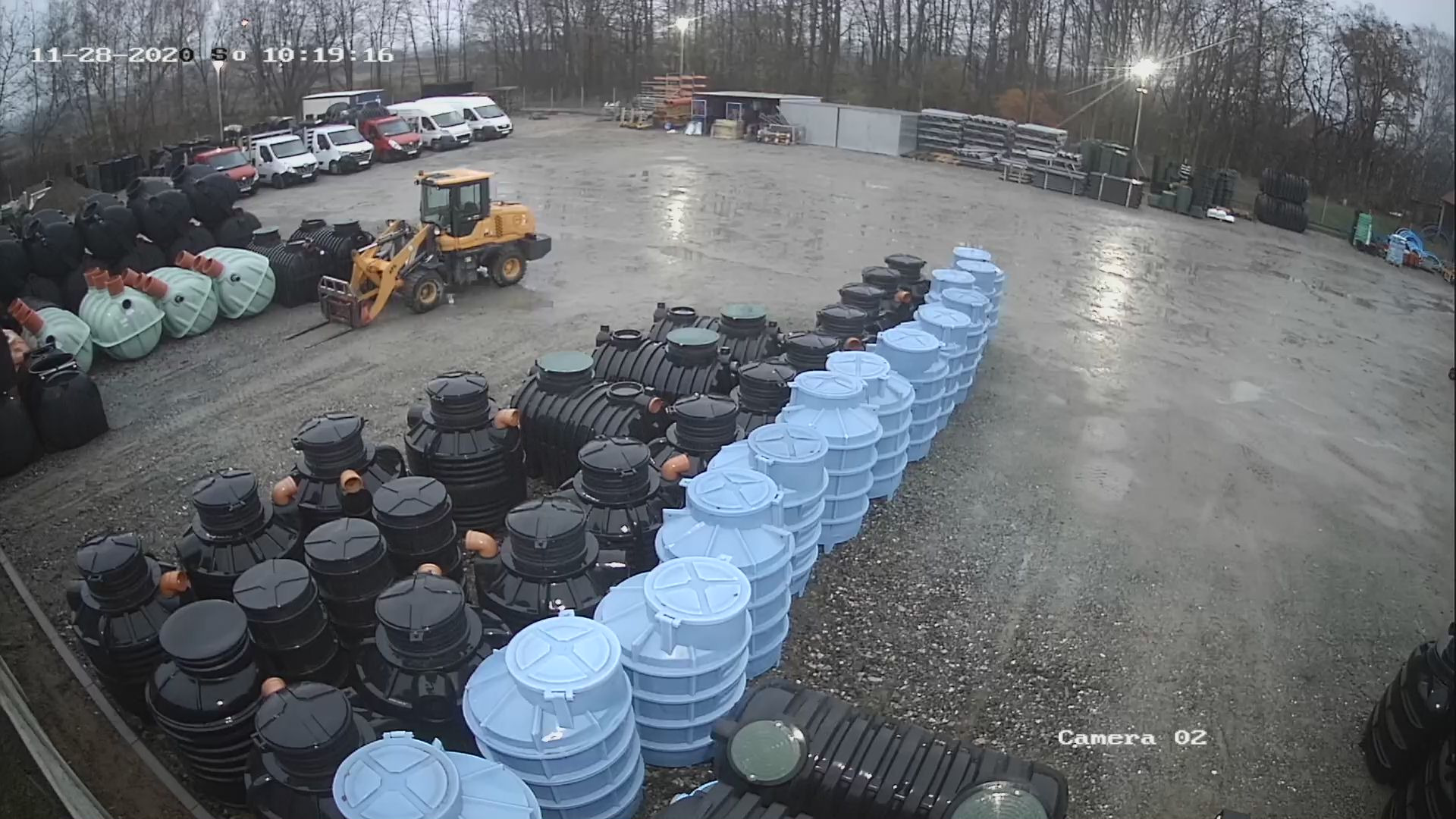 Magazyn zbiorników: rzędy czarnych i błękitnych zbiorników z polietylenu na szarym placu, w tle żółta ładowarka widłowa i zaparkowane samochody dostawcze, u góry data i godzina z kamery przemysłowej.