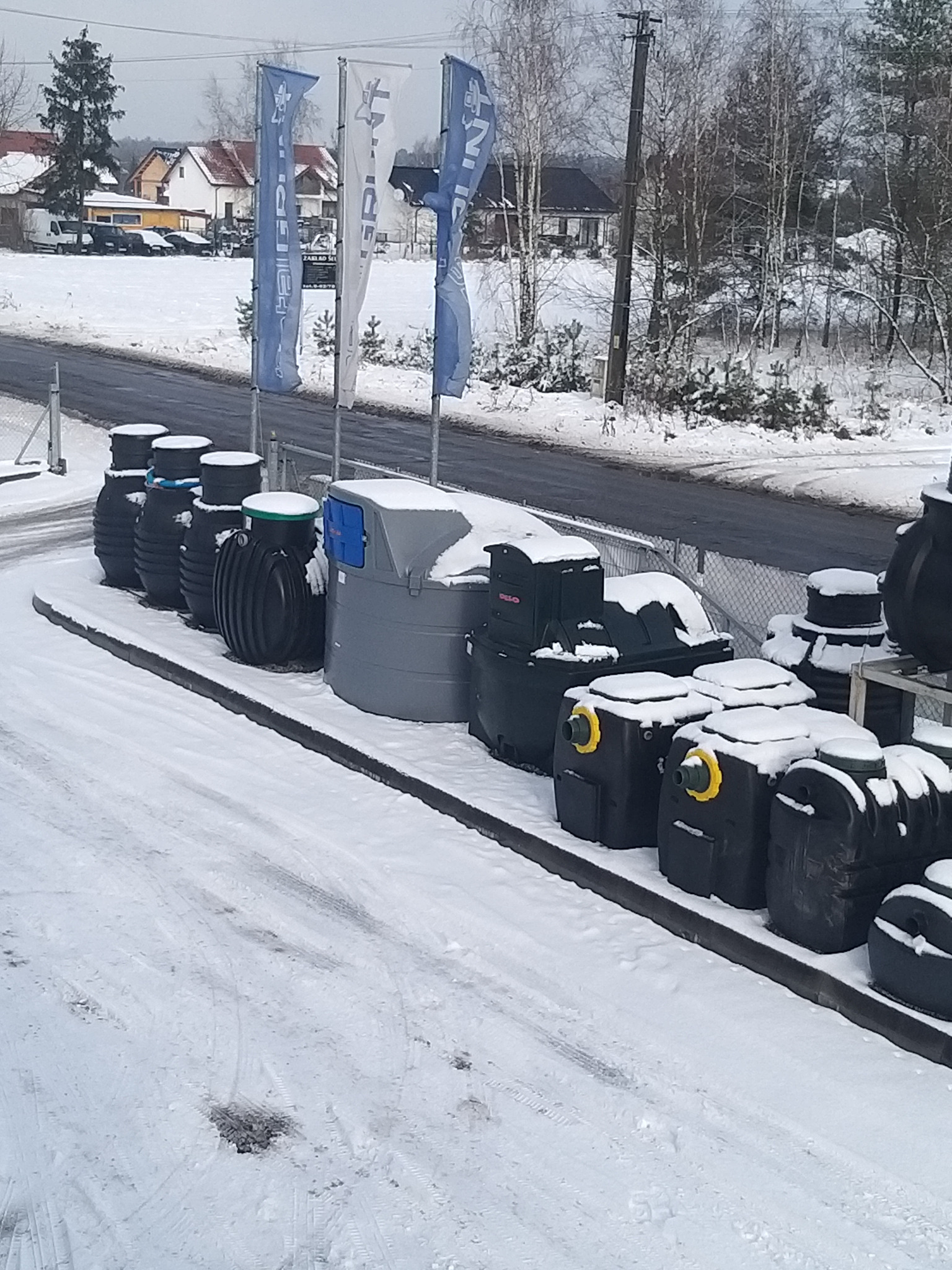Ekspozycja różnych typów przydomowych oczyszczalni ścieków i zbiorników, pokrytych śniegiem, na tle ośnieżonego krajobrazu i flag reklamowych.