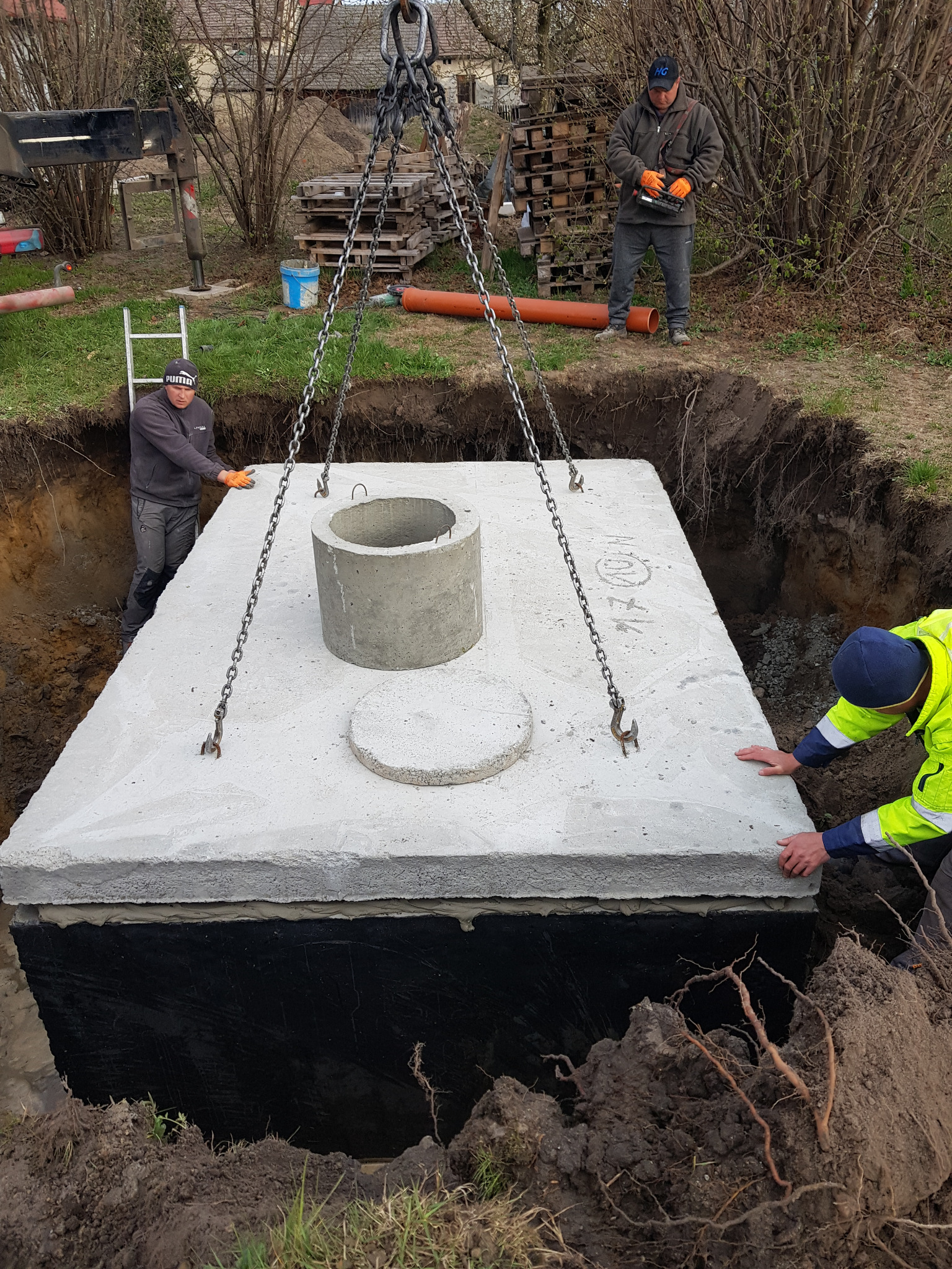 Montaż betonowego szamba w Dziadowej Kłodzie przy użyciu dźwigu; widoczni pracownicy w odzieży roboczej, zabezpieczona czarną folią konstrukcja, teren wykopu.