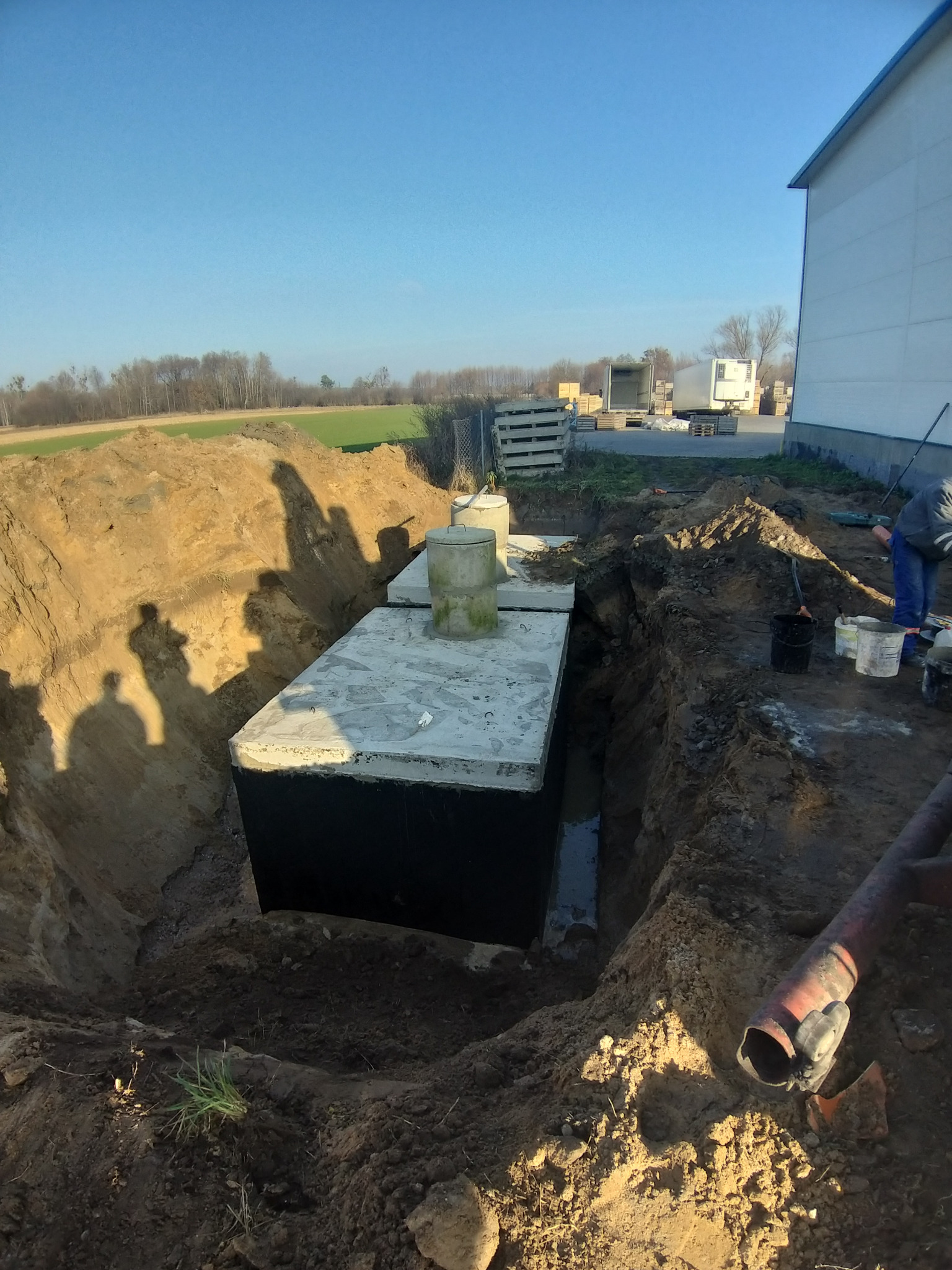 Betonowy zbiornik z dwoma włazami, umieszczony w wykopie ziemnym, częściowo wypełnionym wodą. Na pierwszym planie rura kanalizacyjna. W tle budynek gospodarczy i pole uprawne.