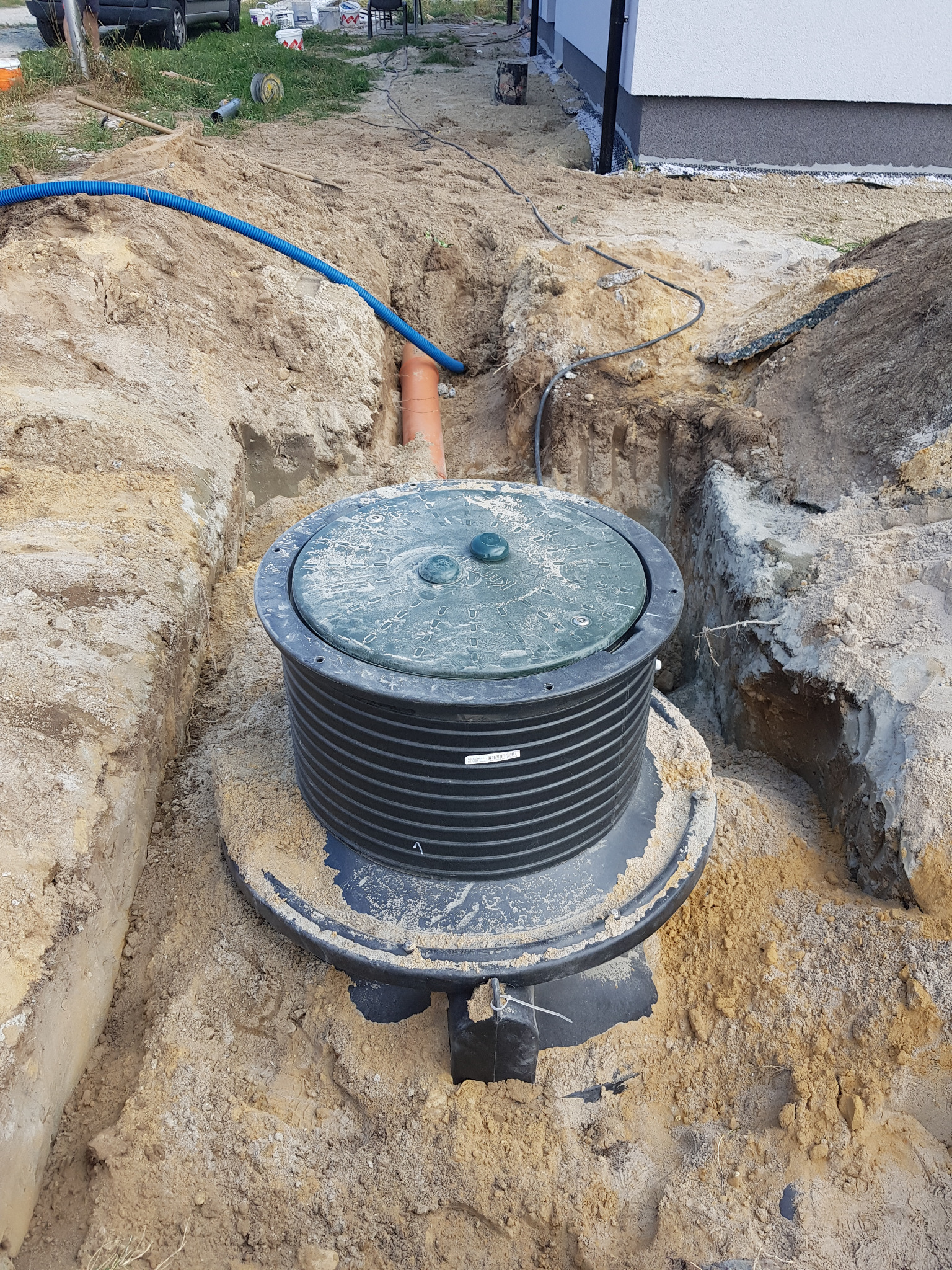 Montaż przydomowej oczyszczalni ścieków: czarny zbiornik z zieloną pokrywą w wykopie, widoczne rury doprowadzające i odprowadzające ścieki, piasek do zasypania, w tle fragment budynku.