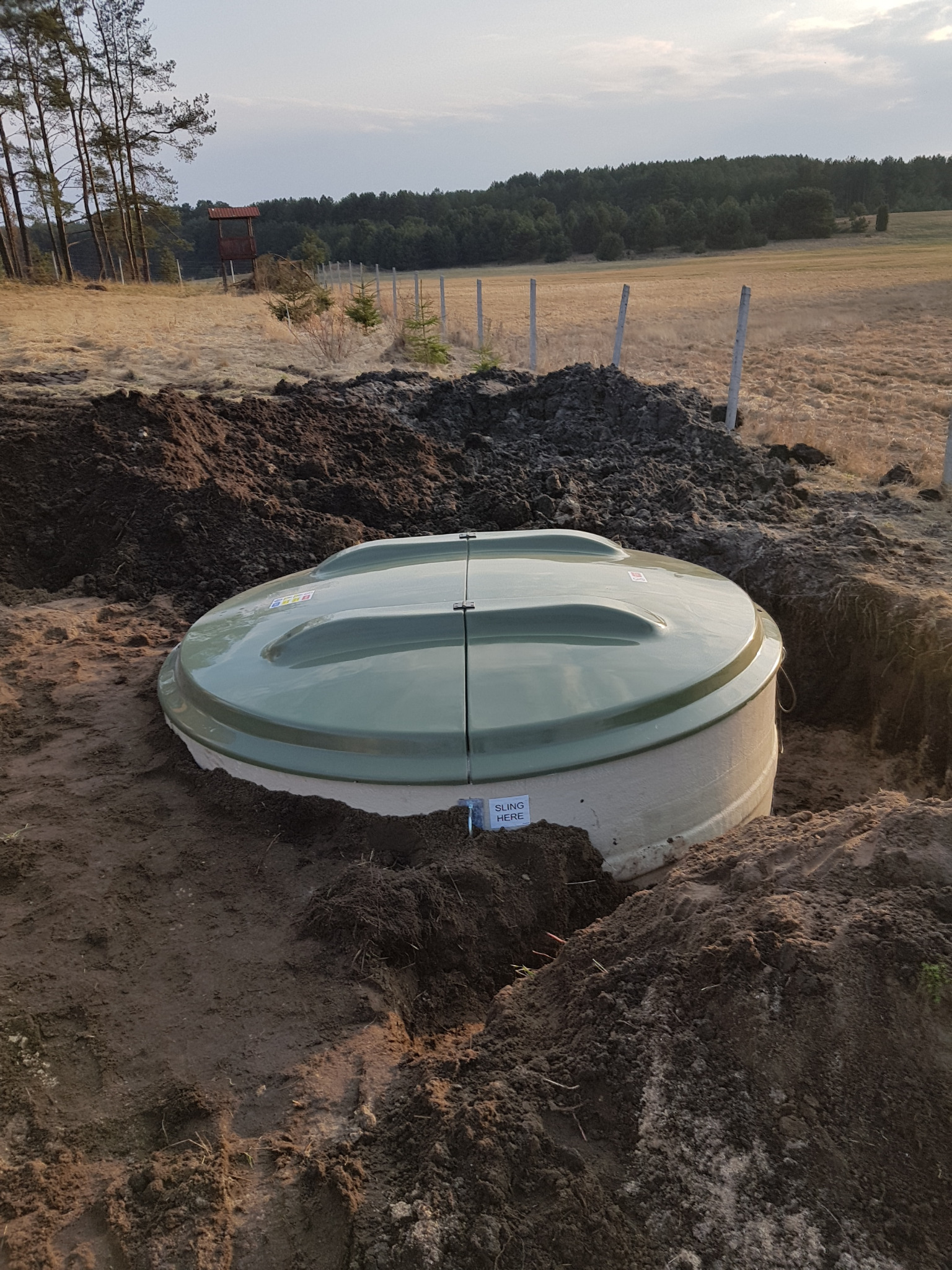 Zielony, okrągły zbiornik z dwudzielną pokrywą umieszczony w wykopie ziemnym na tle łąki i lasu. Widoczna tabliczka z napisem 'SLING HERE'.
