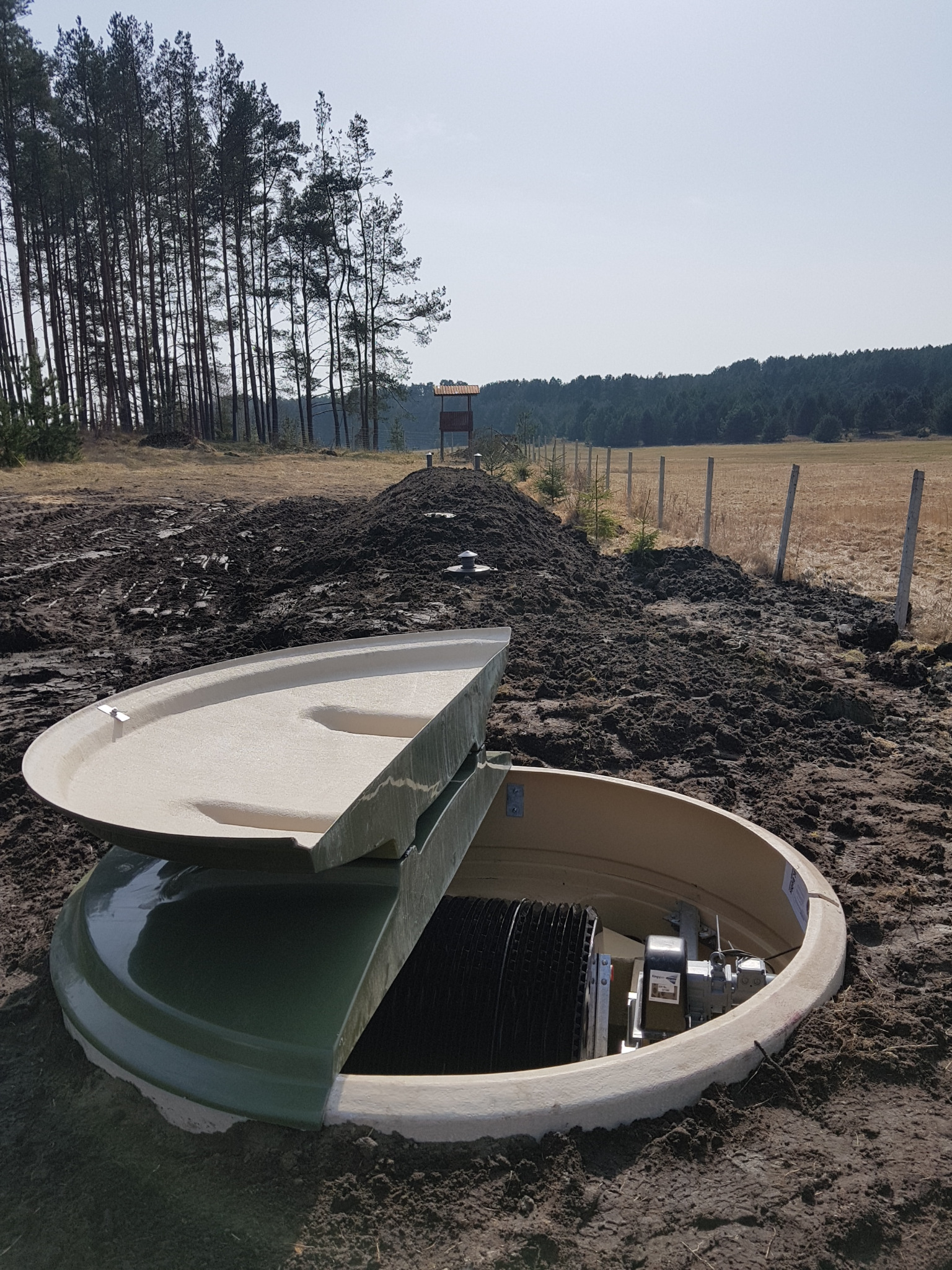 Otwarta właz do betonowego zbiornika z widocznym mechanizmem wewnątrz, umieszczonego w ziemi na tle pola i lasu.