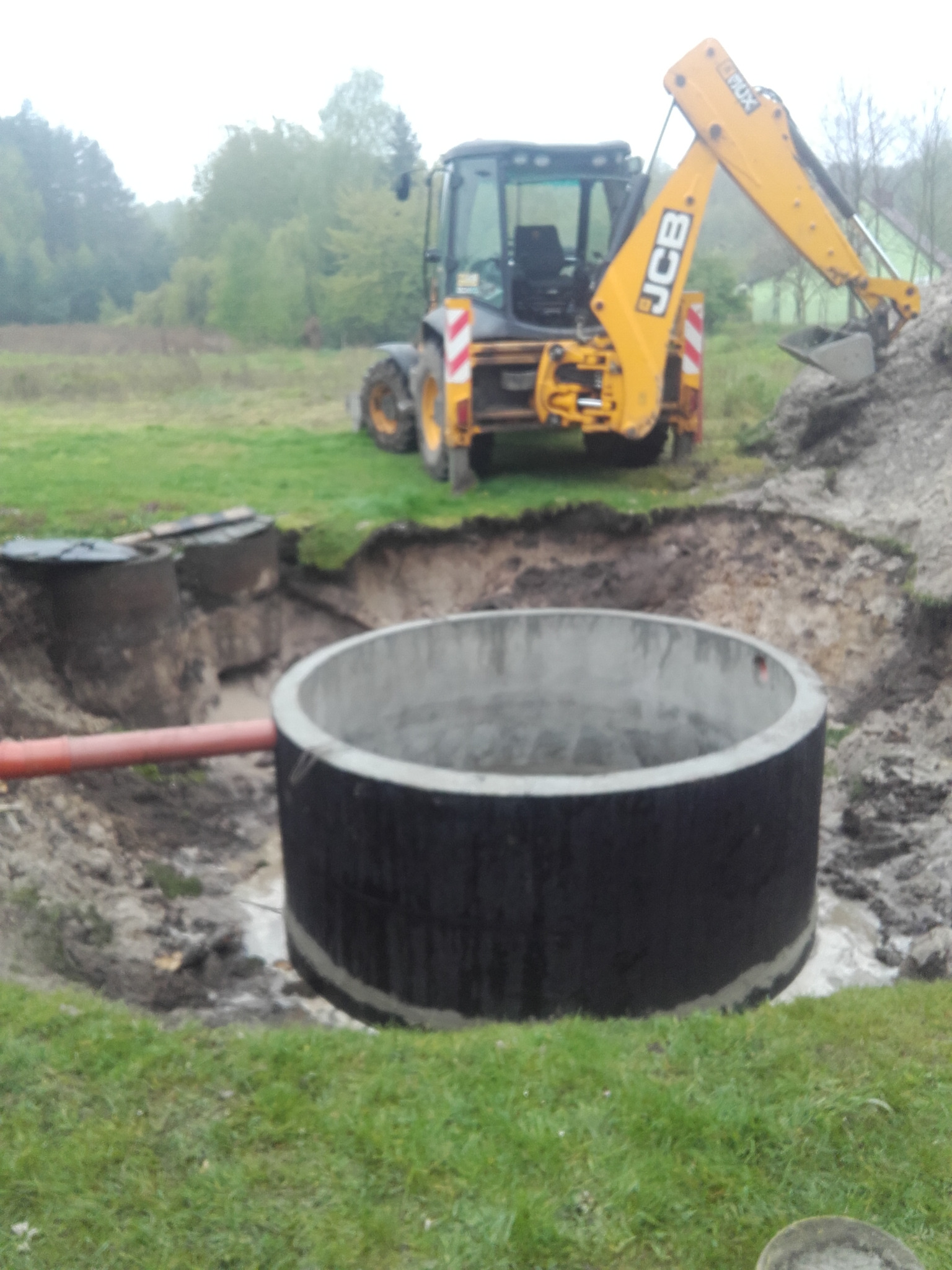 Montaż betonowego kręgu szamba w wykopie, widoczna rura przyłączeniowa i koparka JCB w tle.