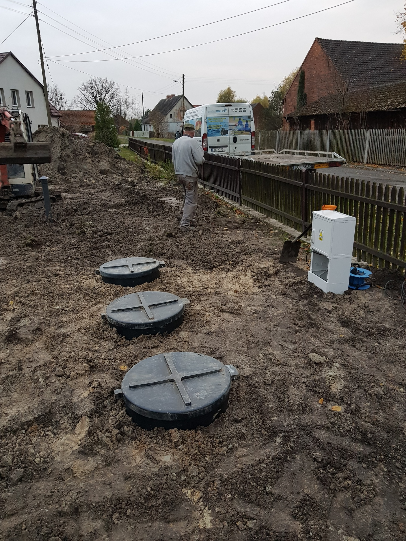 Montaż trzech zbiorników z czarnymi pokrywami na ziemi, obok biała szafka sterownicza, w tle pracownik i samochód dostawczy z reklamą firmy na tle zabudowy wiejskiej.