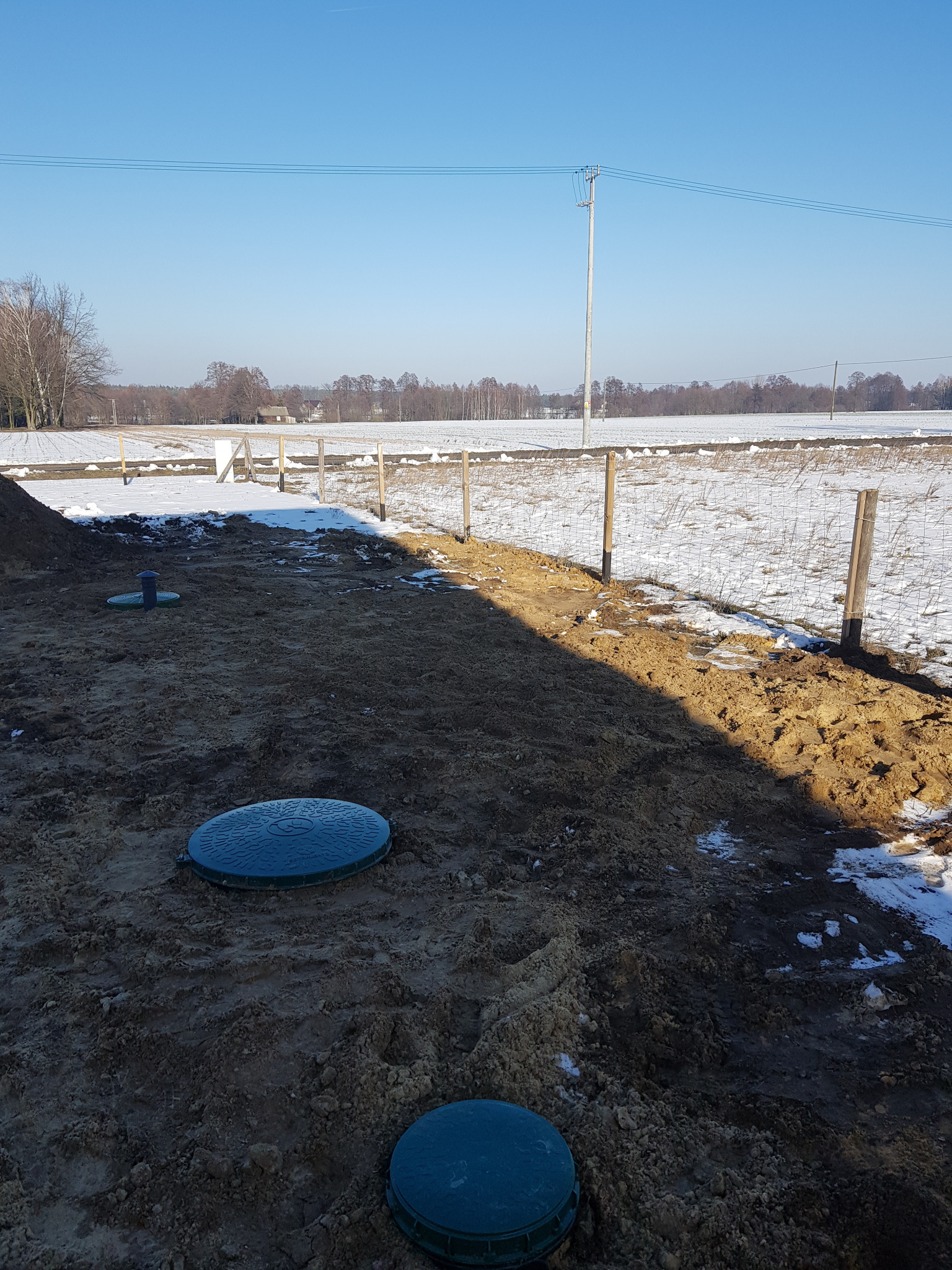 Teren budowy z trzema zielonymi pokrywami włazów kanalizacyjnych w ziemi, częściowo pokrytej śniegiem, w tle pole i linia energetyczna.