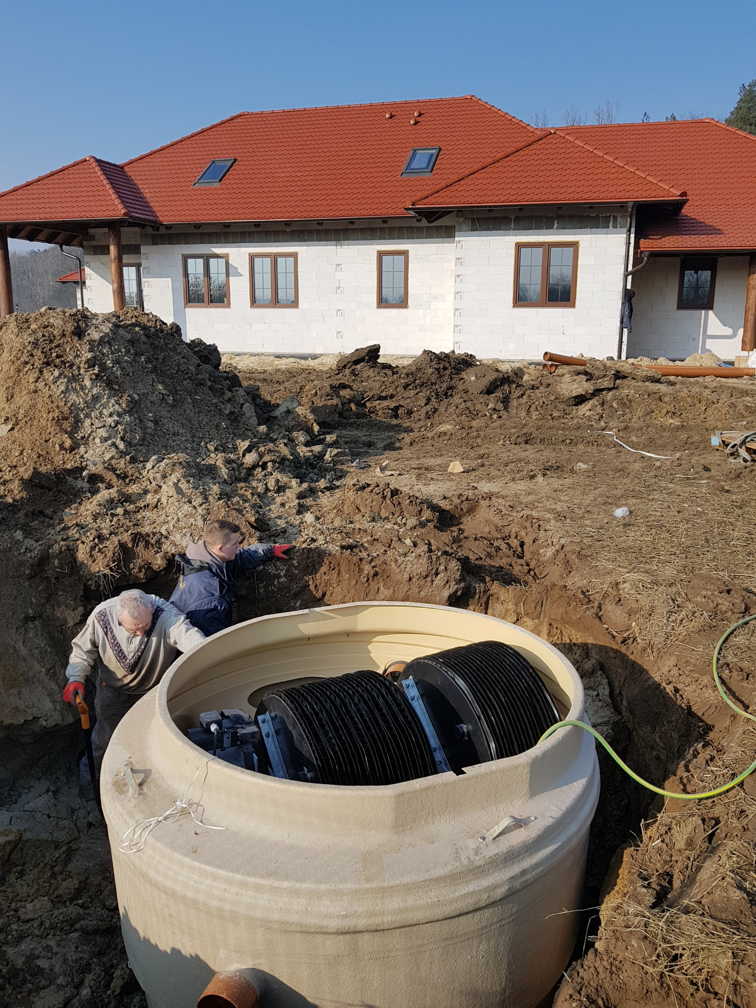 Montaż beżowego zbiornika z czarnymi wkładami w wykopie ziemnym na tle budującego się domu z czerwoną dachówką; dwóch mężczyzn w roboczych ubraniach nadzoruje prace.