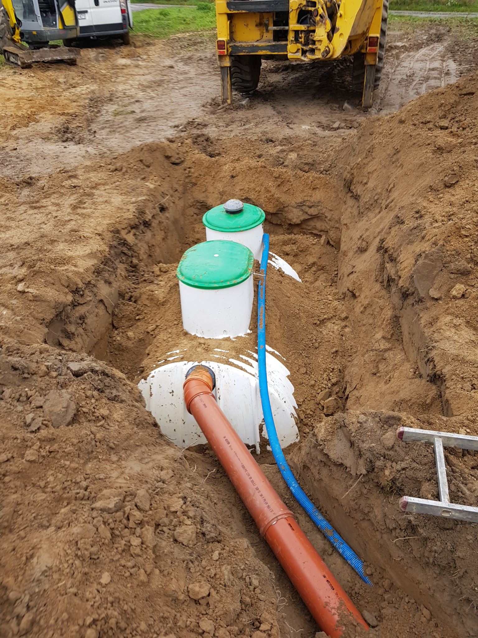 Montaż przydomowej oczyszczalni ścieków w wykopie, widoczne zbiorniki z zielonymi pokrywami, rury kanalizacyjne i maszyny budowlane w tle.