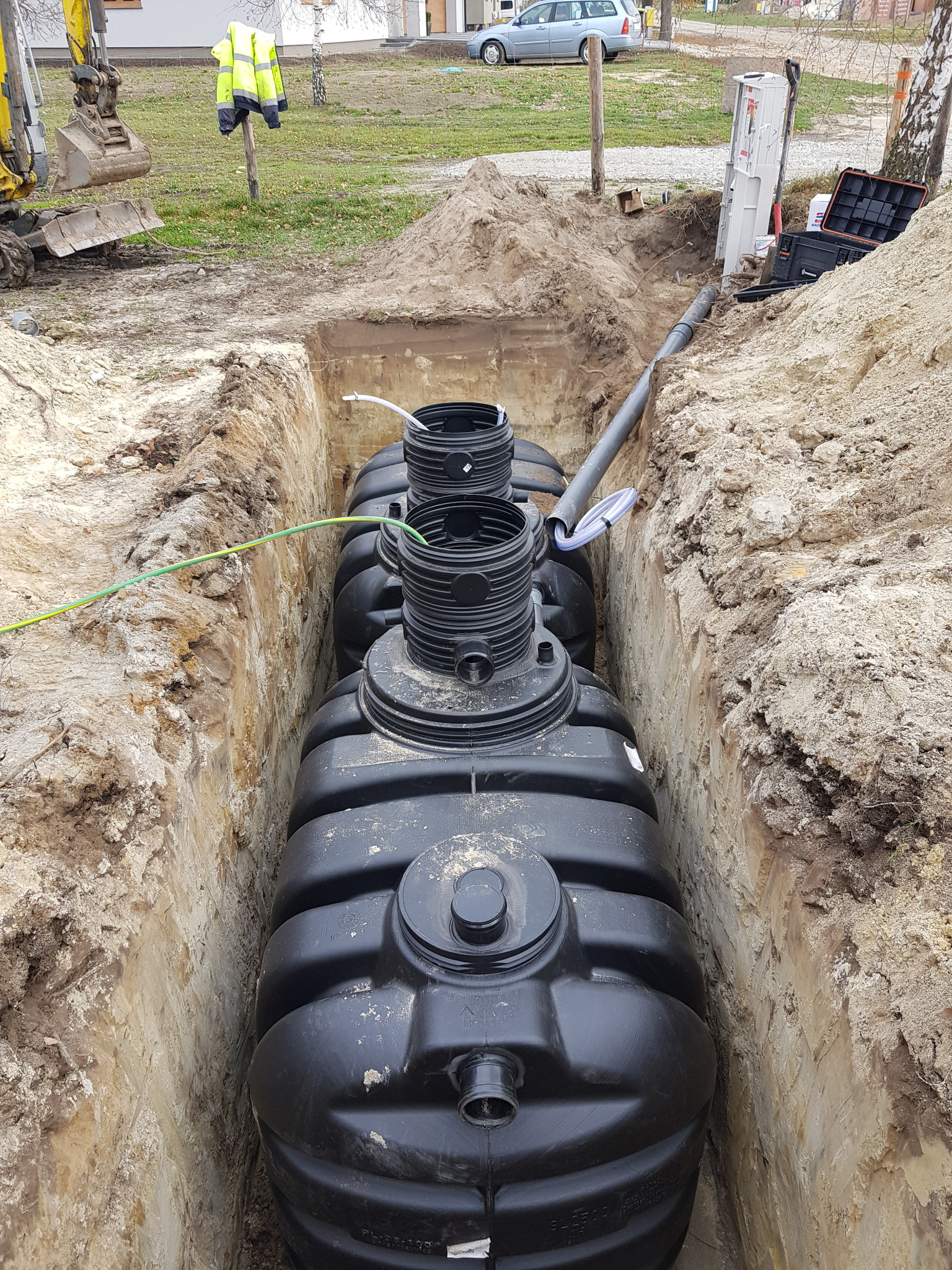 Czarny, zbiornik z tworzywa sztucznego, częściowo zakopany w wykopie ziemnym, widoczne rury i przewody, w tle koparka i budynek mieszkalny.